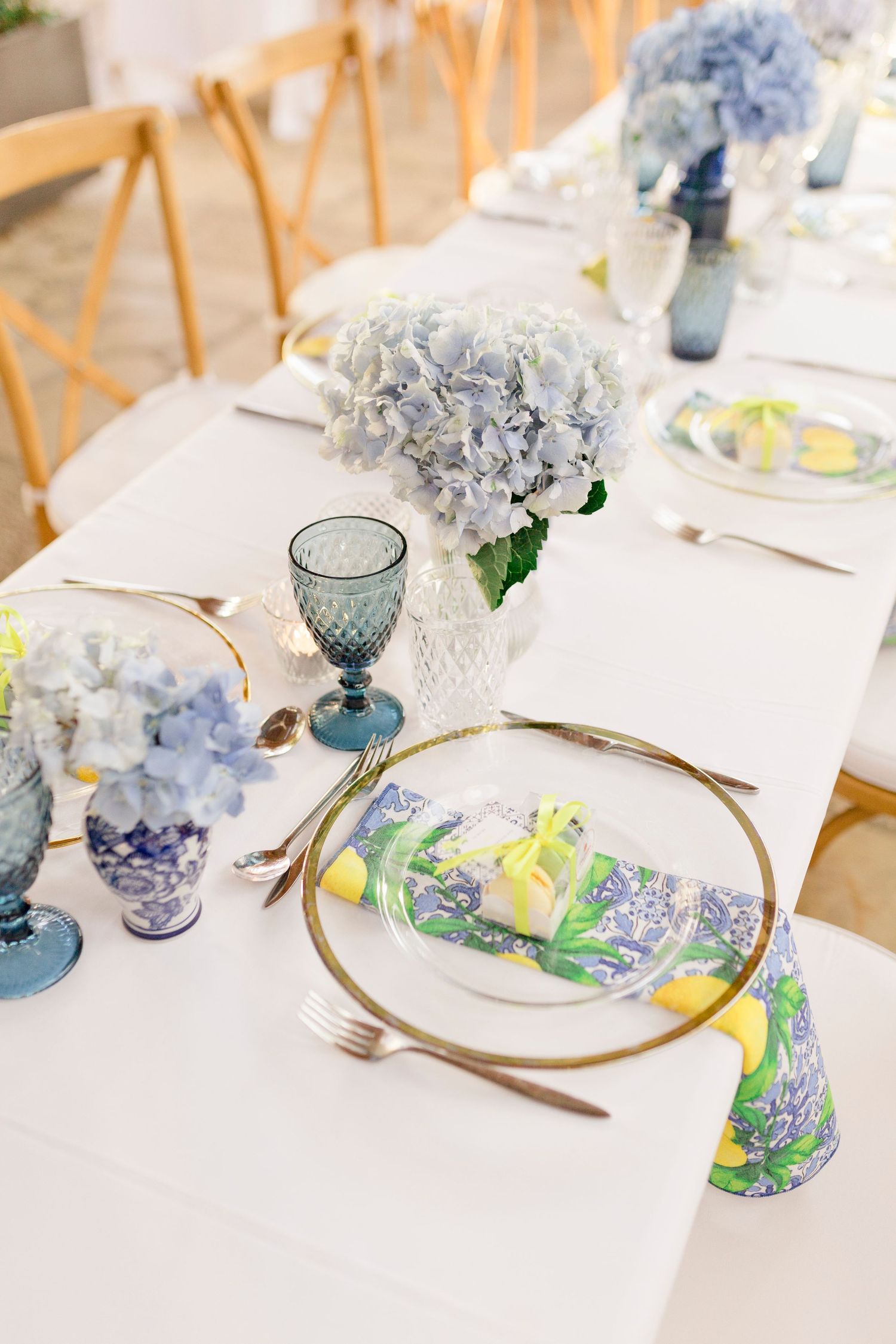 Elegant table setting with blue hydrangeas, gold-rimmed plates, blue goblets, and lemon-print napkins on white linen.