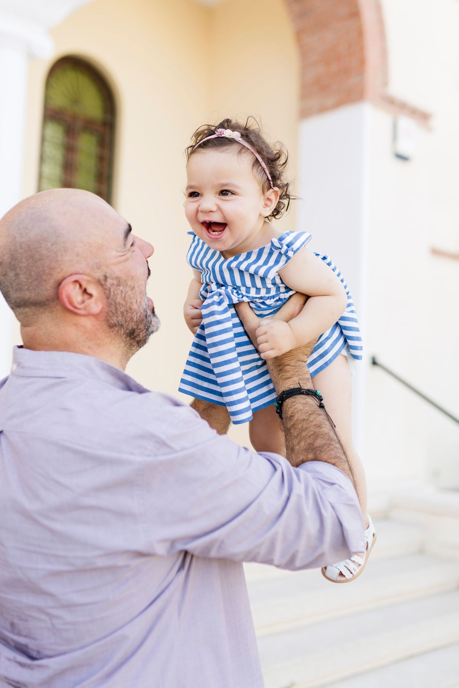 A bald father lifts his laughing baby girl in a blue striped dress outdoors near a building.