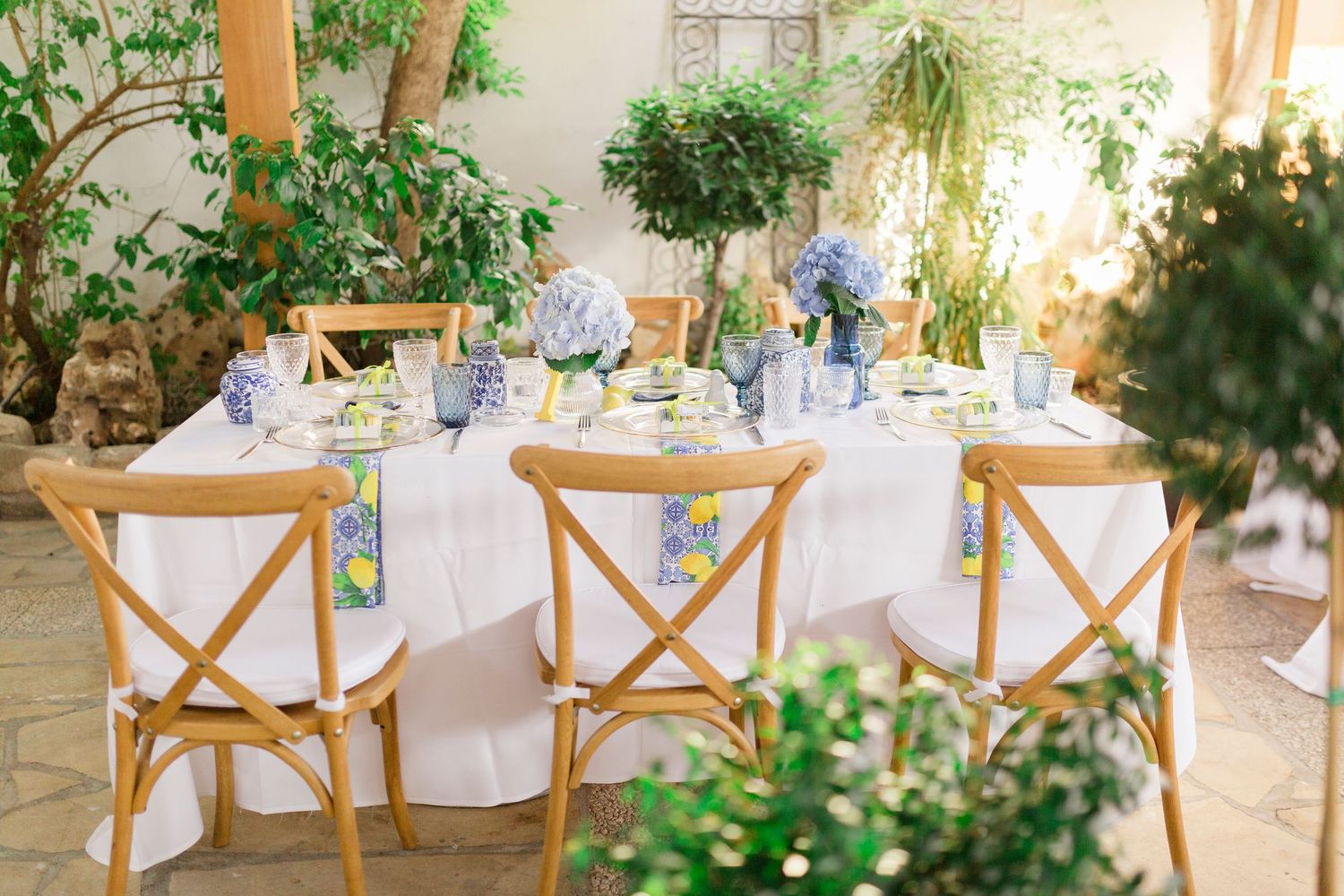 Elegant outdoor wedding reception table with wooden cross-back chairs, white linens, blue floral centerpieces, and lemon accents.