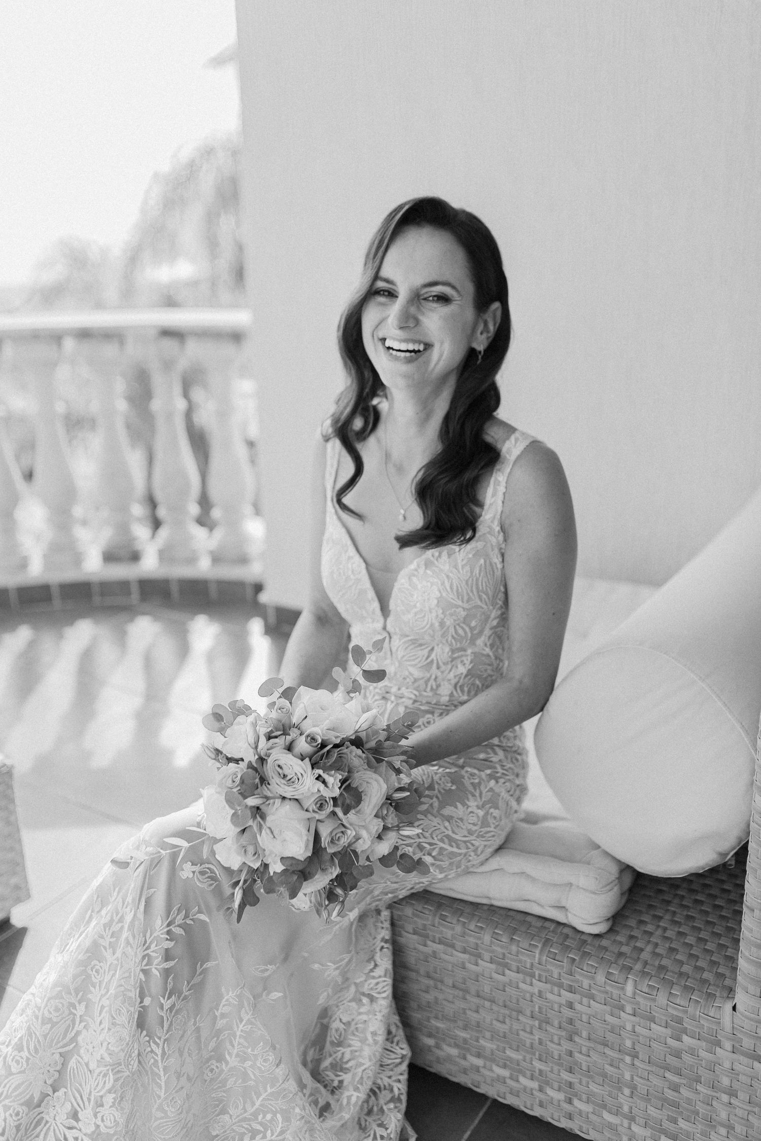 Radiant bride in lace wedding gown laughing while holding bouquet on balcony in black and white portrait.