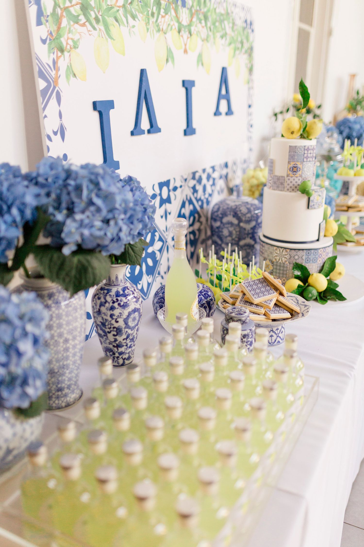 Blue and white Greek-themed dessert table with hydrangeas, tiered cake, lemon decor, and ΙΛΙΑ letters backdrop.