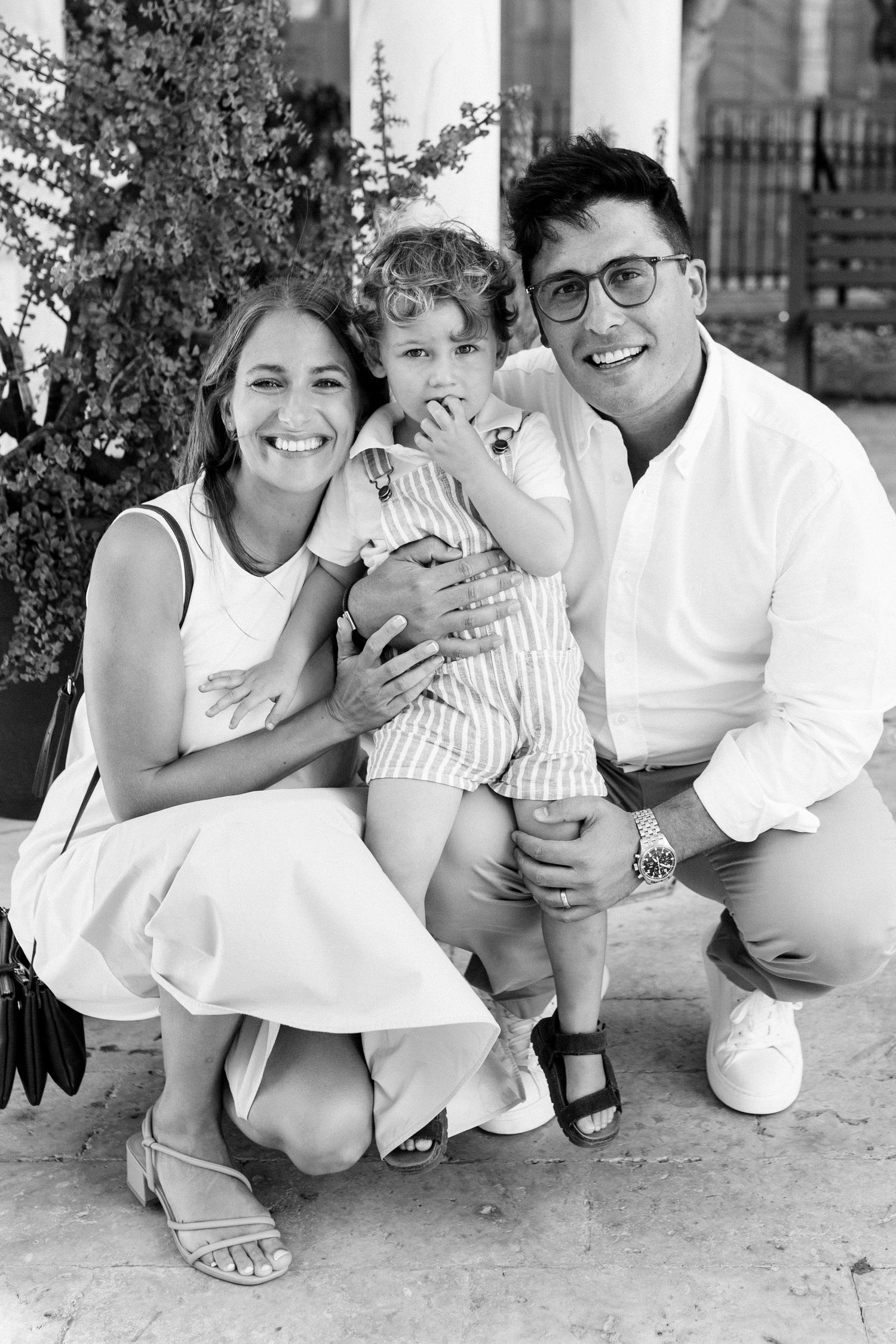 Black and white photo of a smiling family with a toddler posing outdoors near columns and greenery.