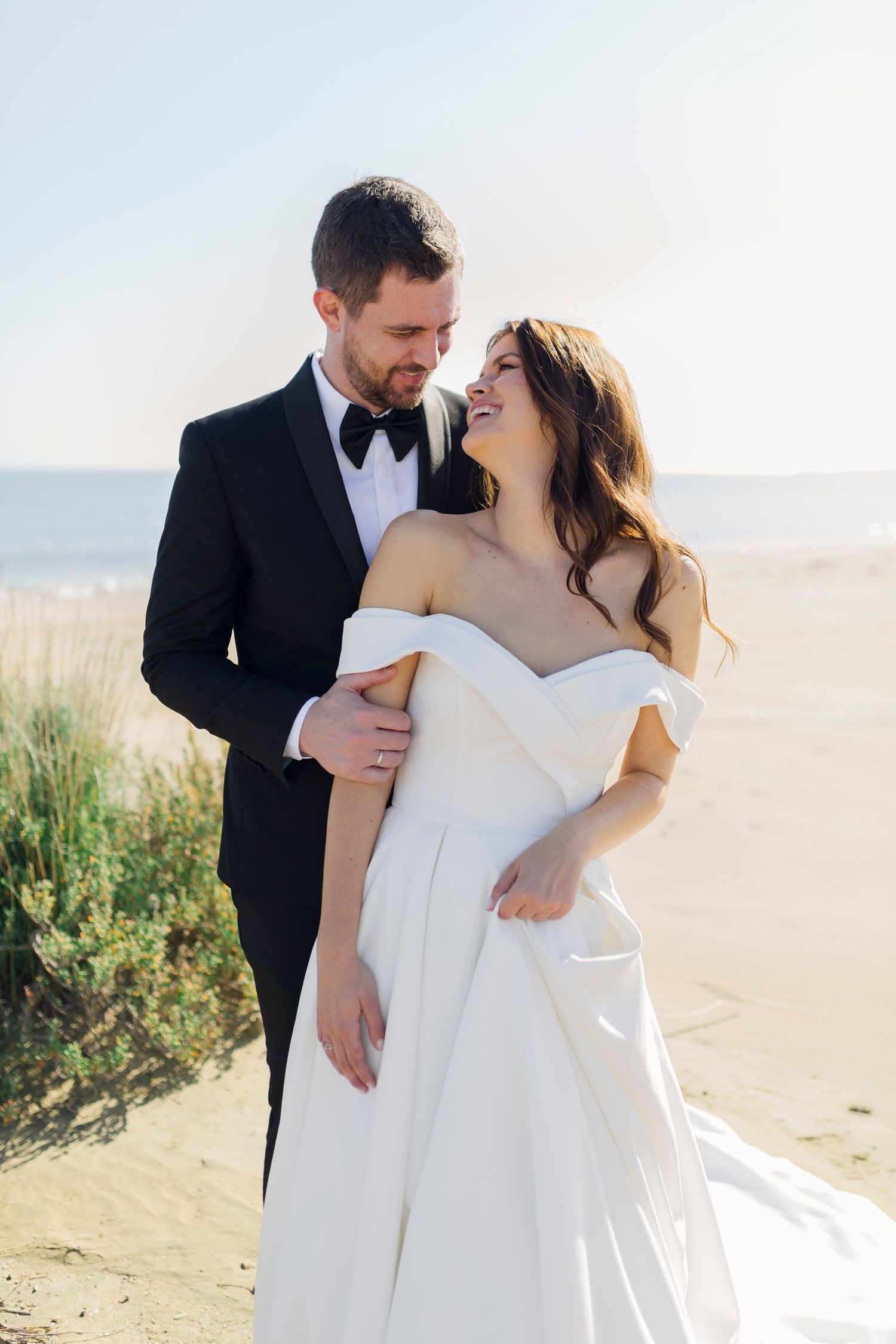 A smiling bride in an off-shoulder white gown embraces her groom in a black tuxedo on a sunny beach.
