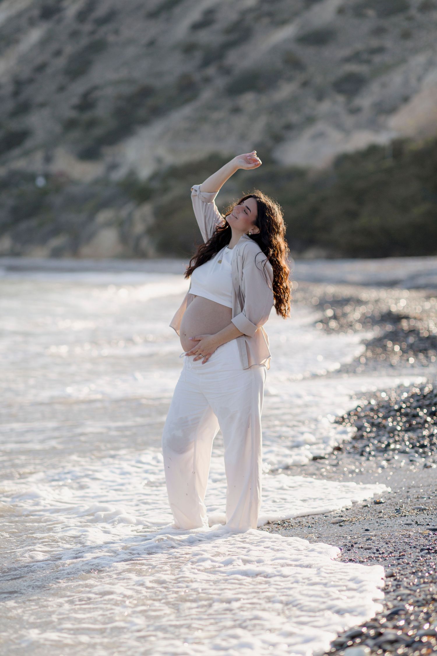 Pregnant woman in white outfit standing in ocean waves on a pebble beach, cradling her belly with hills behind.
