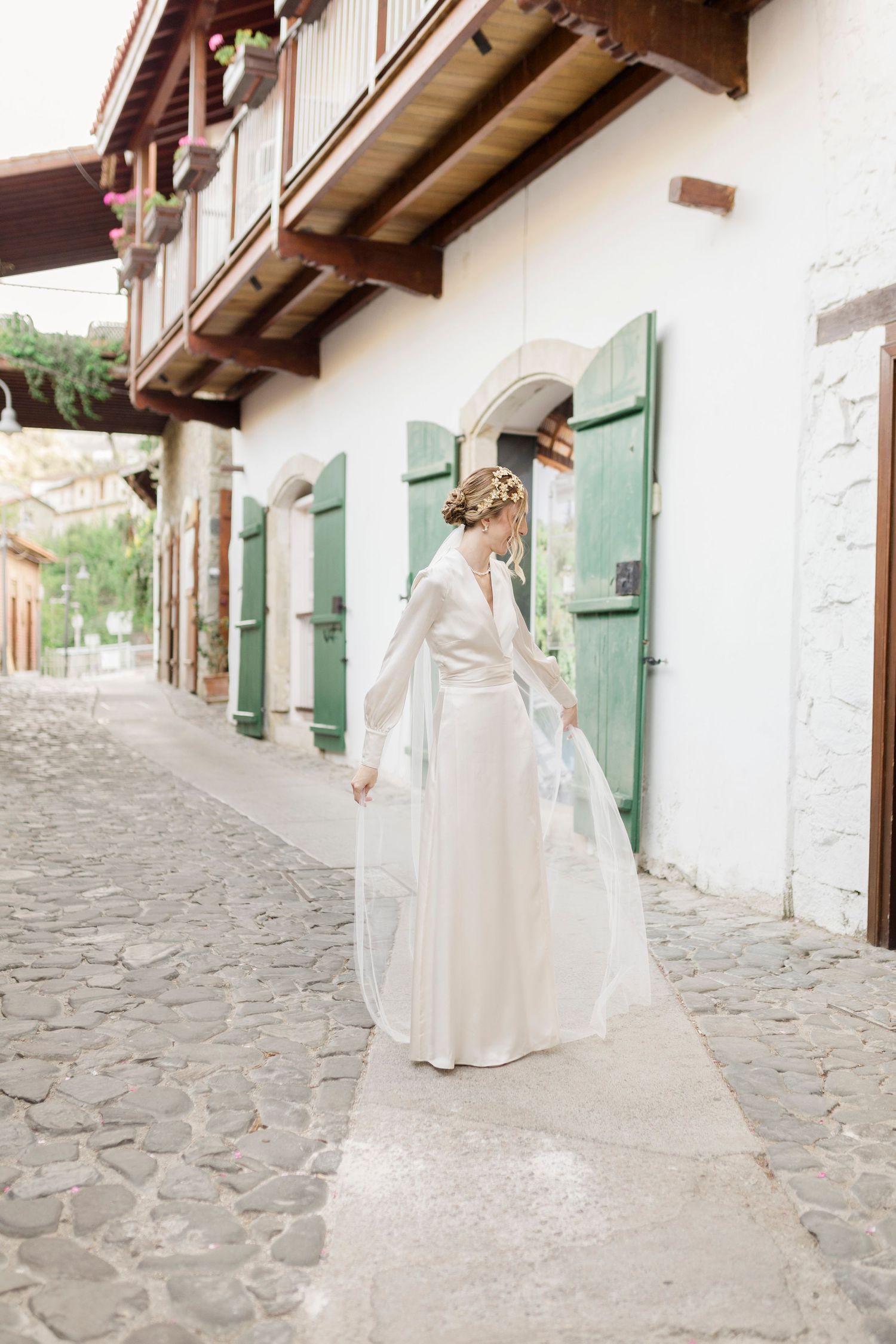 Bride in elegant white gown with veil walks cobblestone street past white building with green shutters.