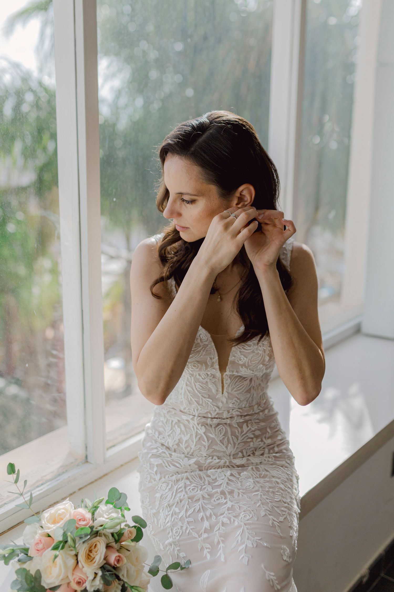Bride in lace wedding gown putting on earrings by a window with a floral bouquet nearby.