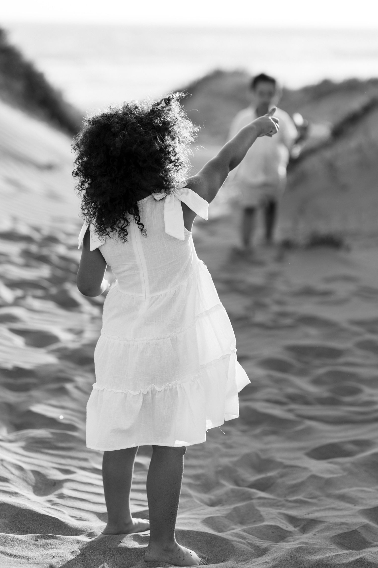 Black and white photo of a curly-haired girl in white dress reaching toward a person on sandy beach dunes.