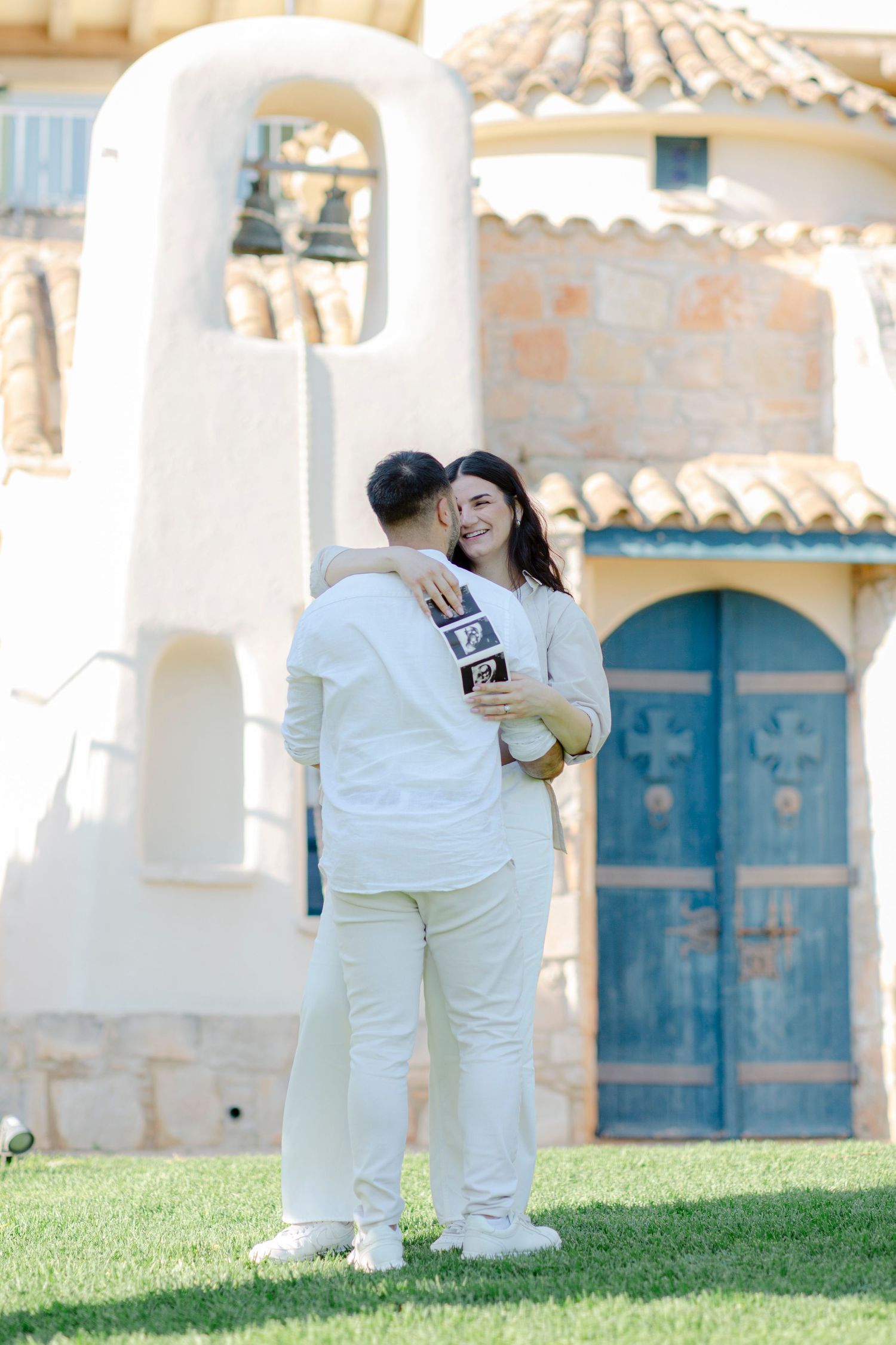 Couple in white outfits embracing outdoors, holding ultrasound photos near a rustic building with a blue arched door.