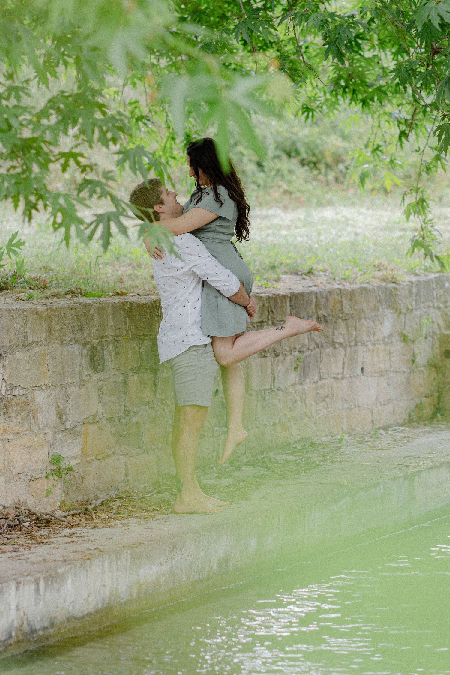 Couple sharing a romantic embrace near a stone wall by a green pool surrounded by trees.