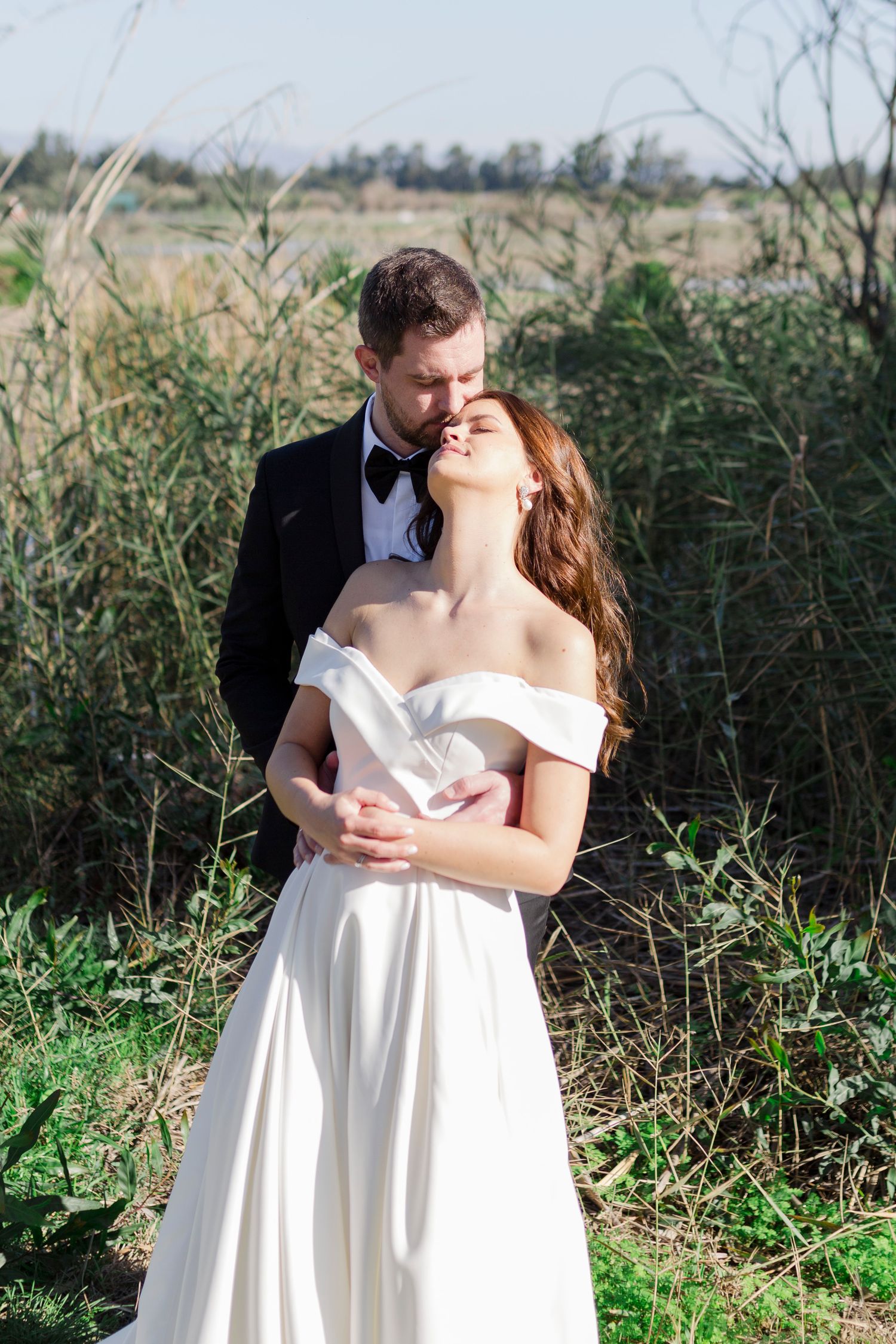 Bride and groom embracing outdoors in nature, bride in off-shoulder white gown, groom in black tuxedo with bow tie.