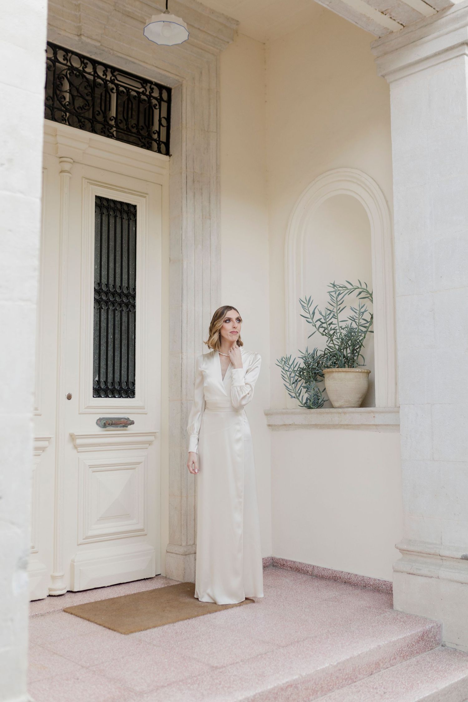 Elegant bride in long white satin dress standing at classical building entrance.