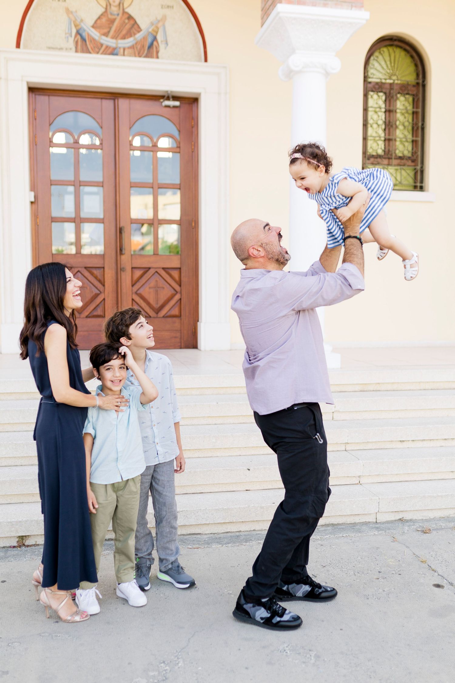 Family of five outside a church, father lifting toddler in the air while mother and two sons watch and smile.
