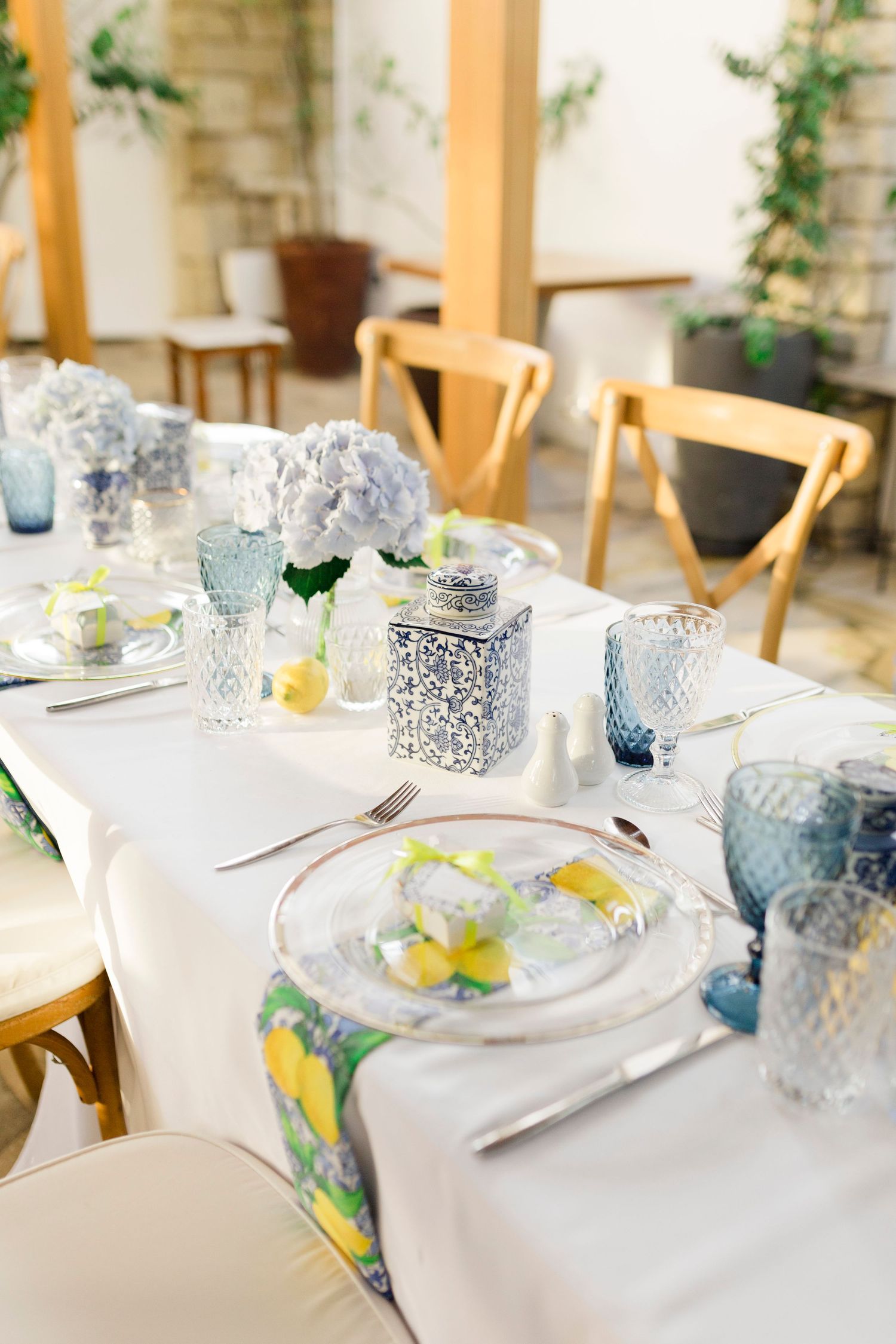 Elegant wedding table setting with blue hydrangeas, lemons, blue glassware, and white linens in a bright venue.