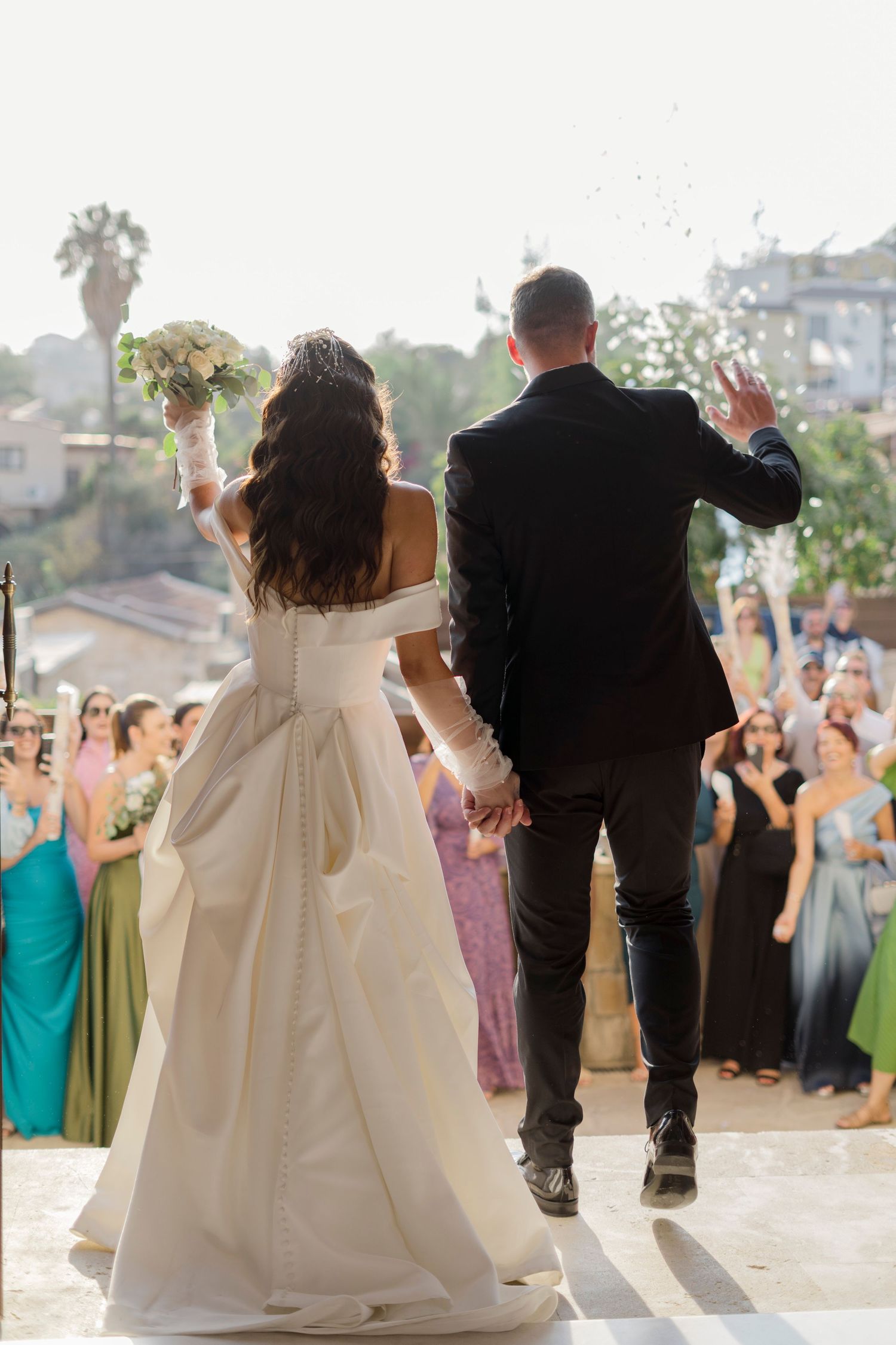 Bride in white gown and groom in black suit wave to guests as they exit their outdoor wedding ceremony.