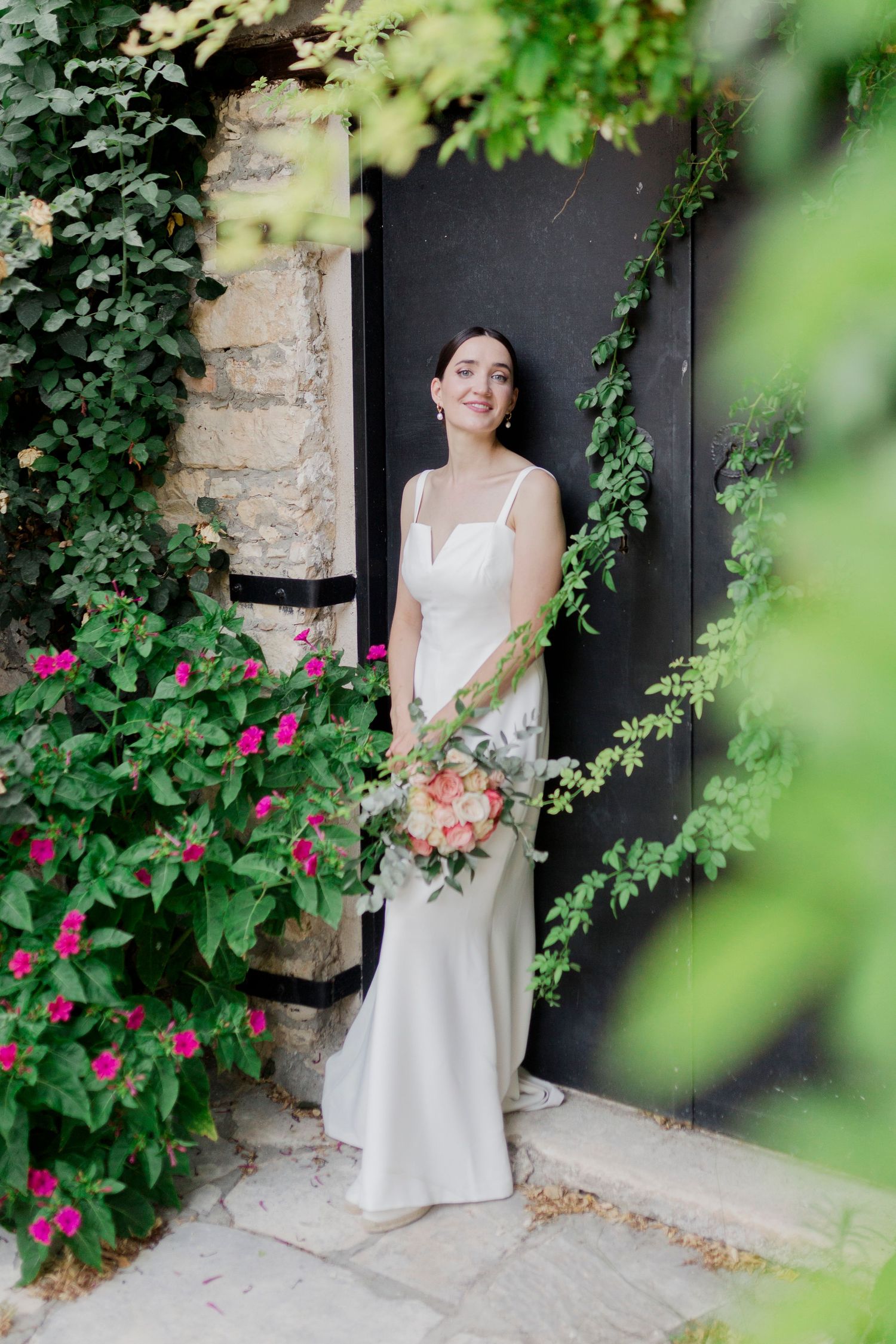 Smiling bride in elegant white slip gown holding peach bouquet standing in doorway surrounded by green ivy.
