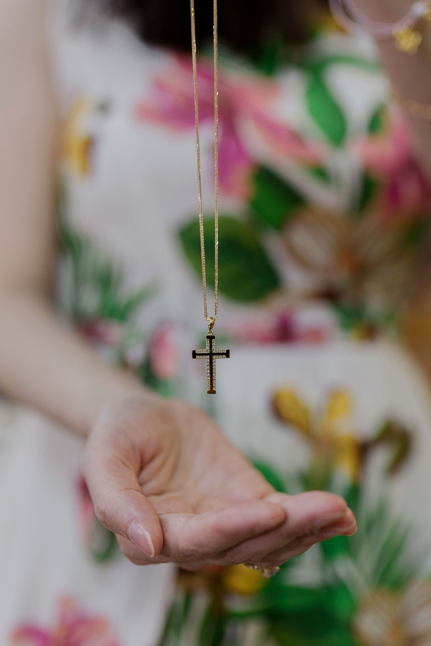 Woman in floral dress holding a gold cross necklace pendant in her outstretched hand.