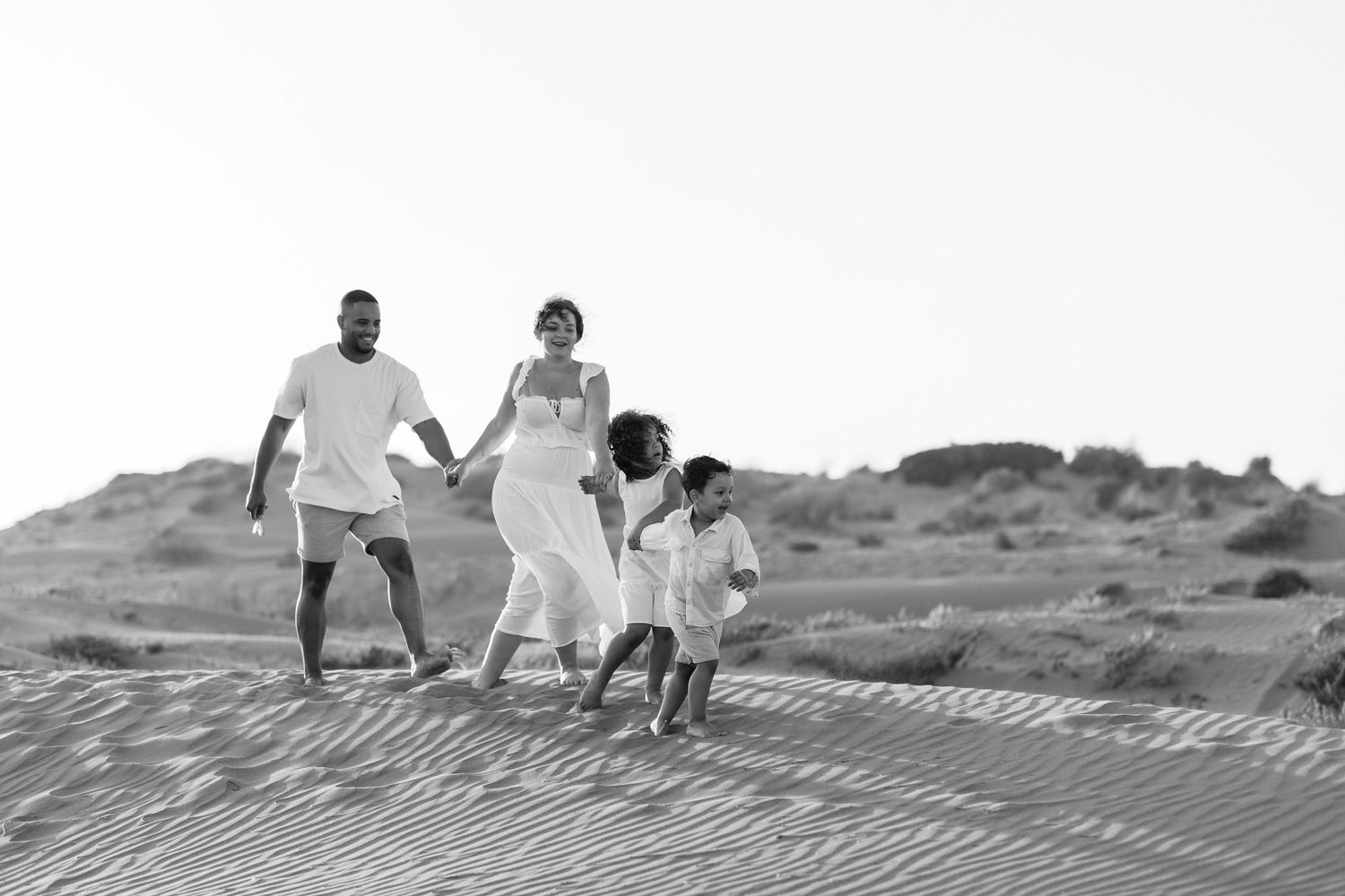 Black and white photo of a family of four walking on sand dunes, parents holding children's hands at the beach.