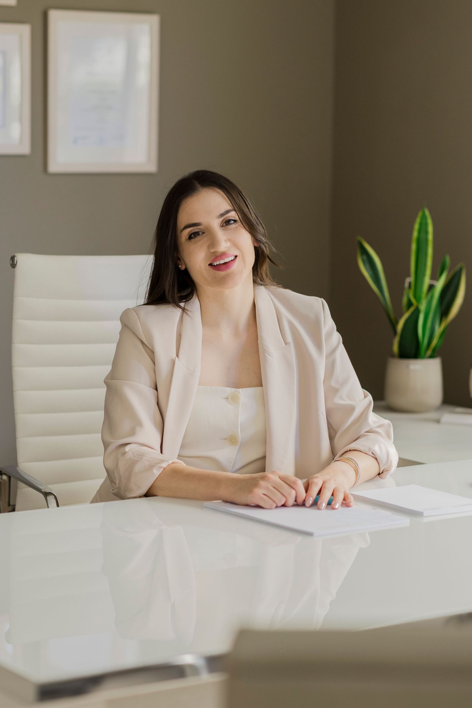 Professional woman in beige blazer sitting at white desk in modern office with plant and framed art.