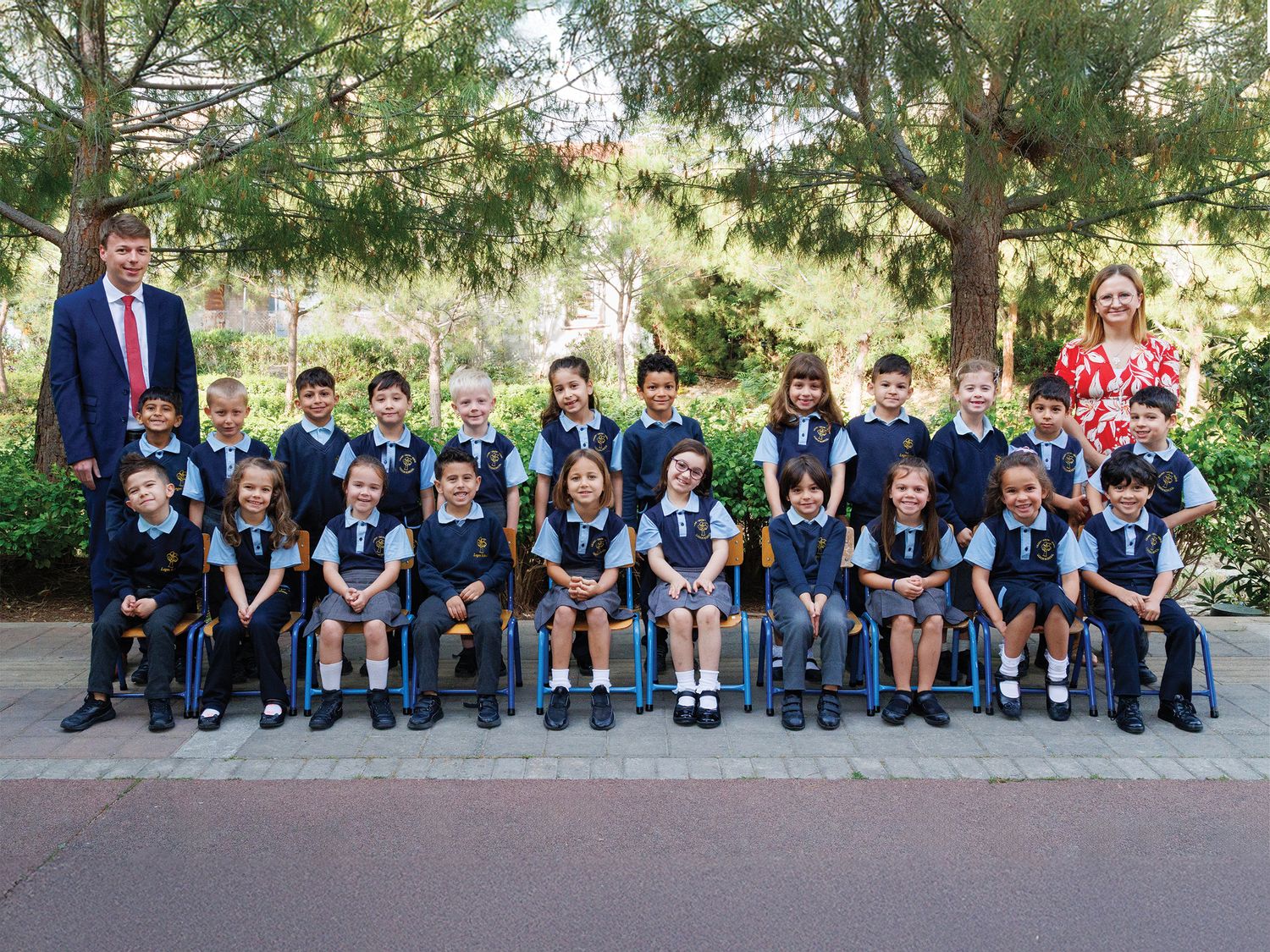 School class photo of young students in uniforms posing with two teachers outdoors among green trees.