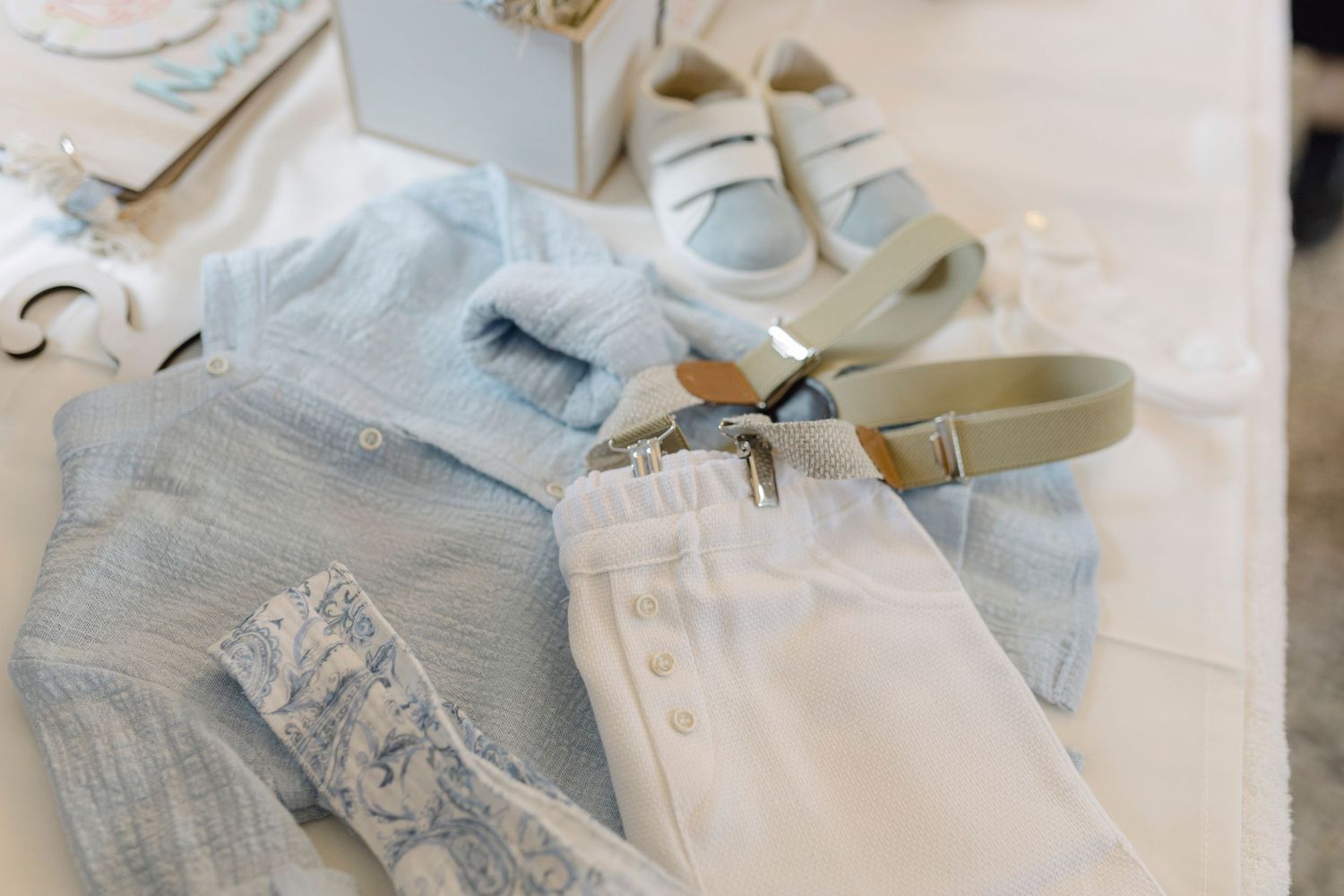 Baby boy baptism outfit with light blue shirt, white pants, suspenders, and white sneakers on a table.