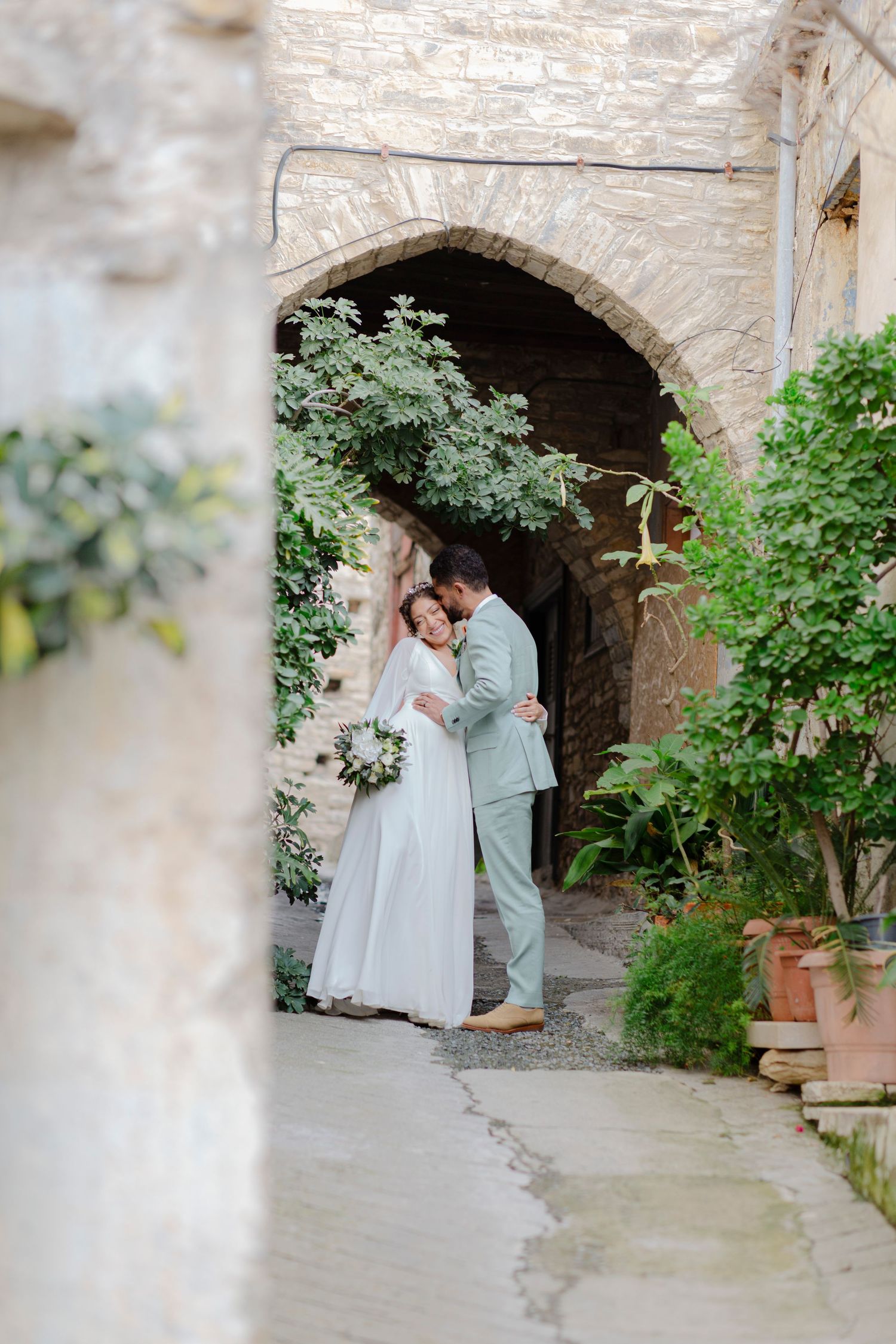 Bride and groom embracing under stone archway surrounded by lush greenery at elegant outdoor wedding venue.