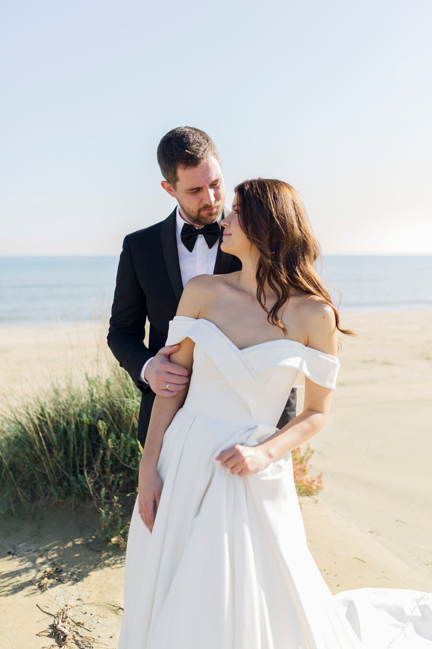 Bride in off-shoulder white gown and groom in black tuxedo share romantic moment on sandy beach.