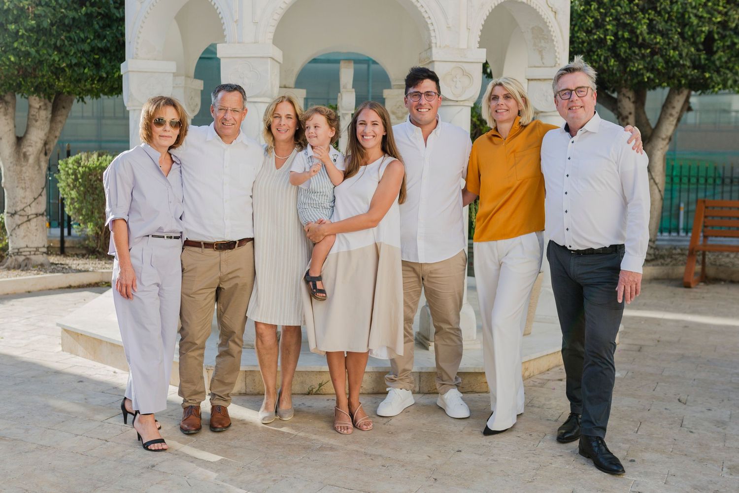 Group of seven adults posing together outdoors near white arched architecture in a sunny Mediterranean setting.