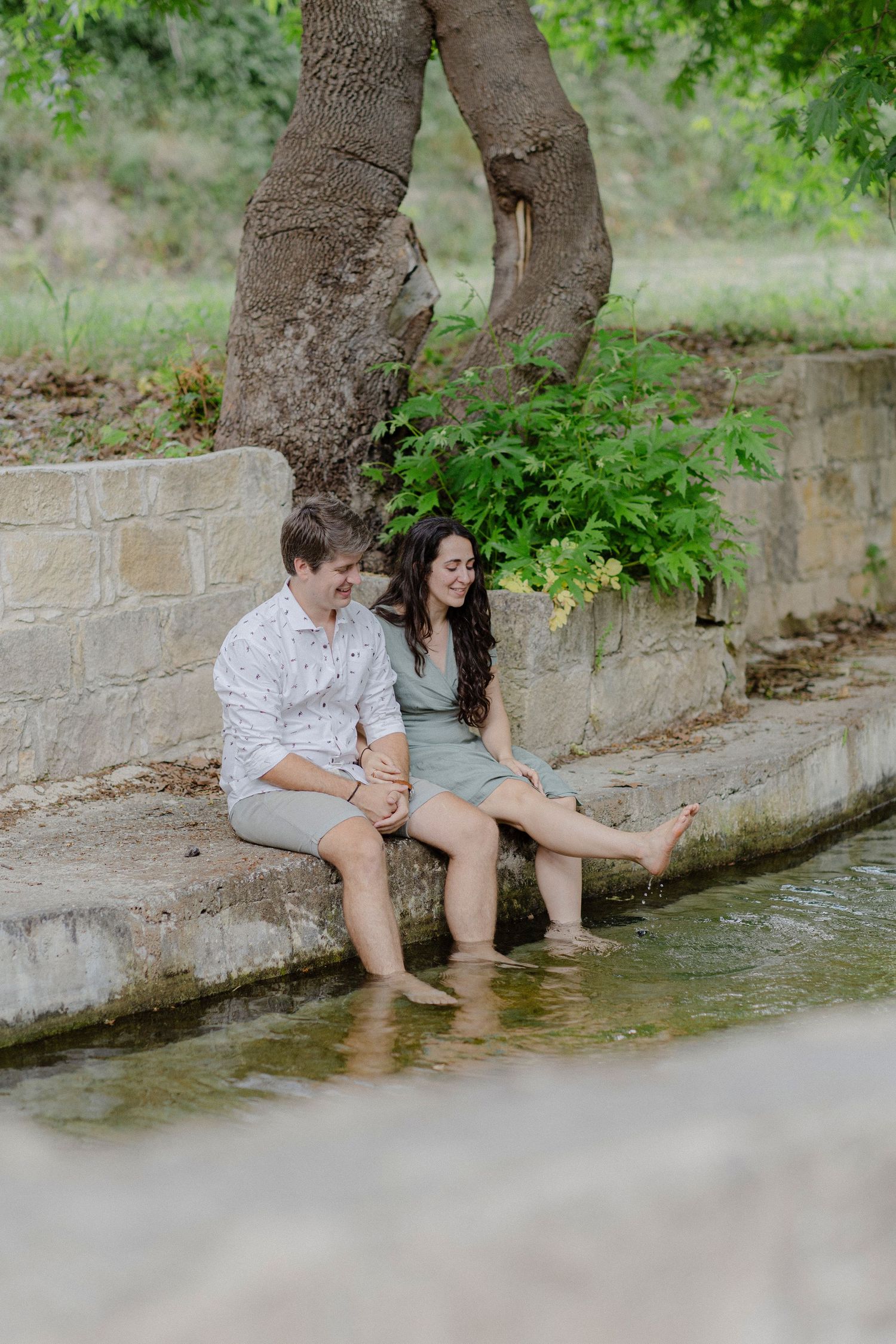 A couple sits barefoot by a stone water feature, laughing together under a large tree in a lush park.