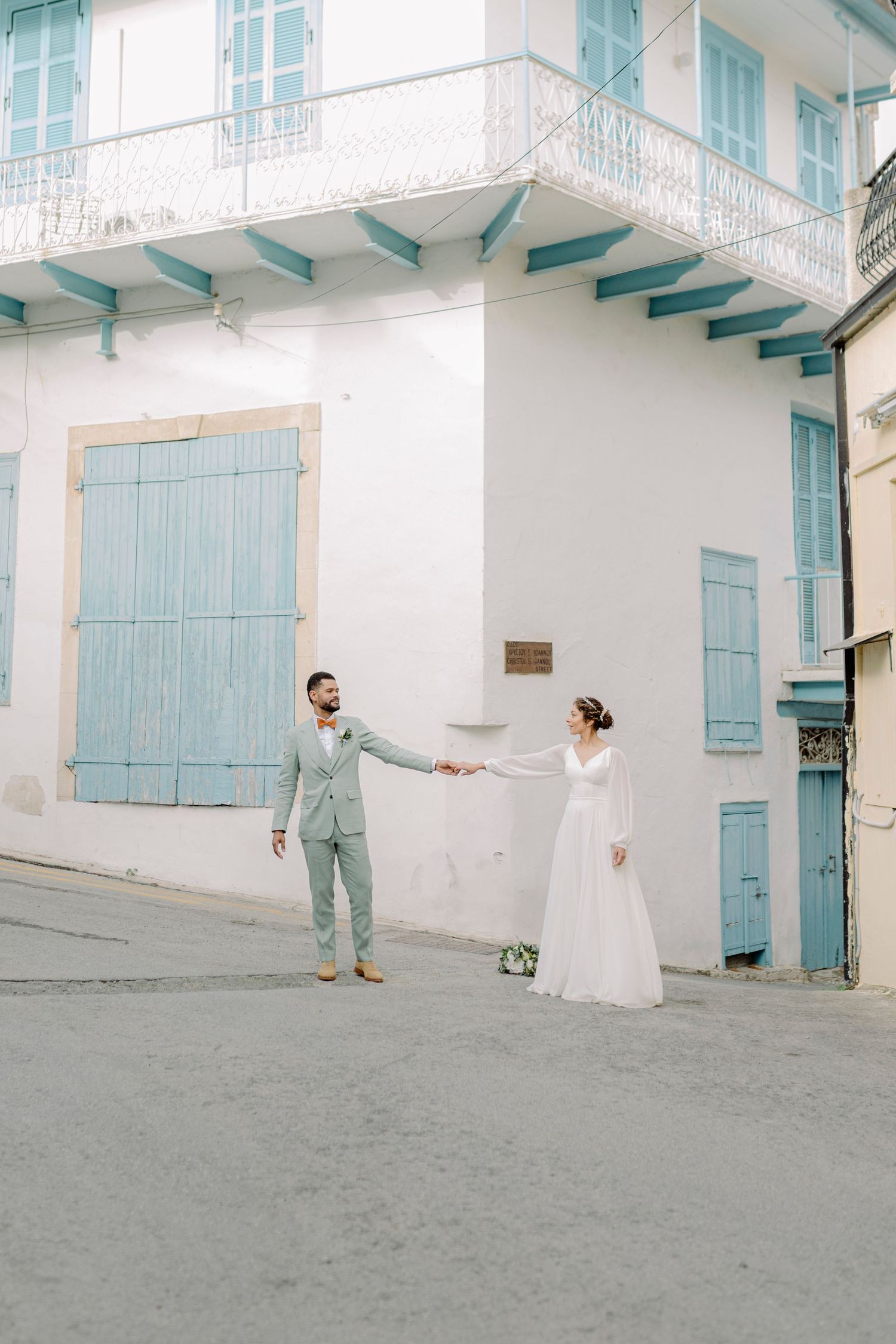 Bride and groom hold hands in front of a white building with blue shutters on their wedding day.