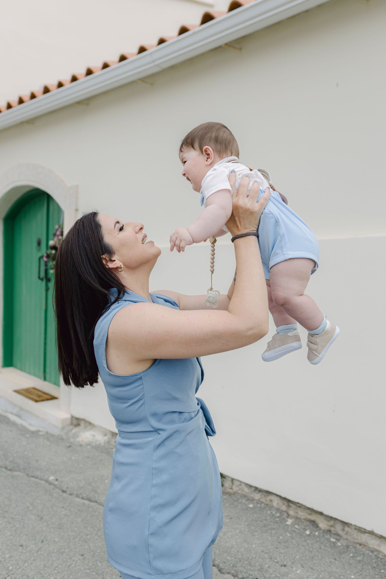 A woman in a blue dress joyfully lifts a smiling baby in the air outdoors near a white building.