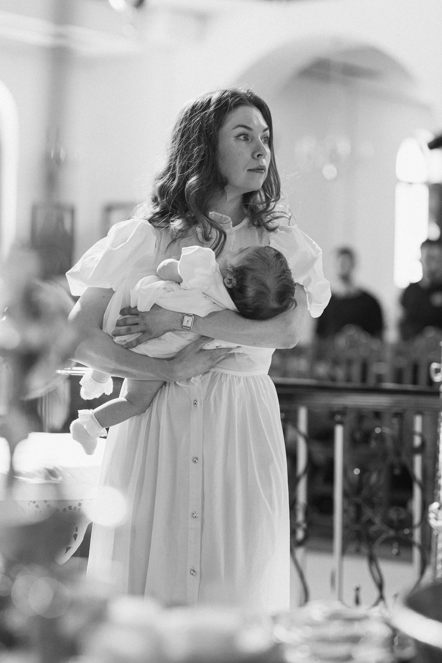 Woman in white dress holds sleeping newborn during a church baptism ceremony in black and white.