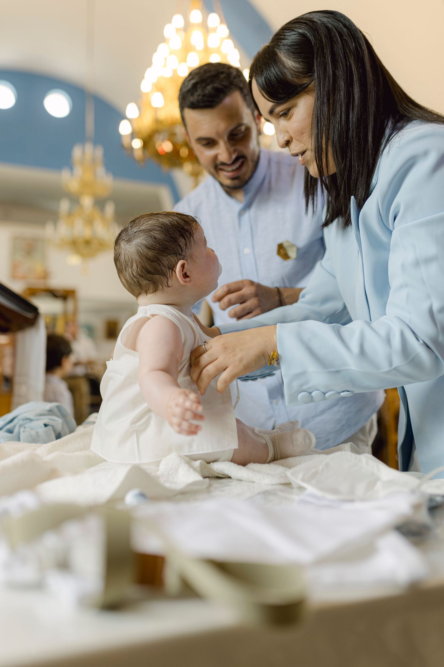 Parents dressing baby in white christening gown at church baptism ceremony with chandelier backdrop.
