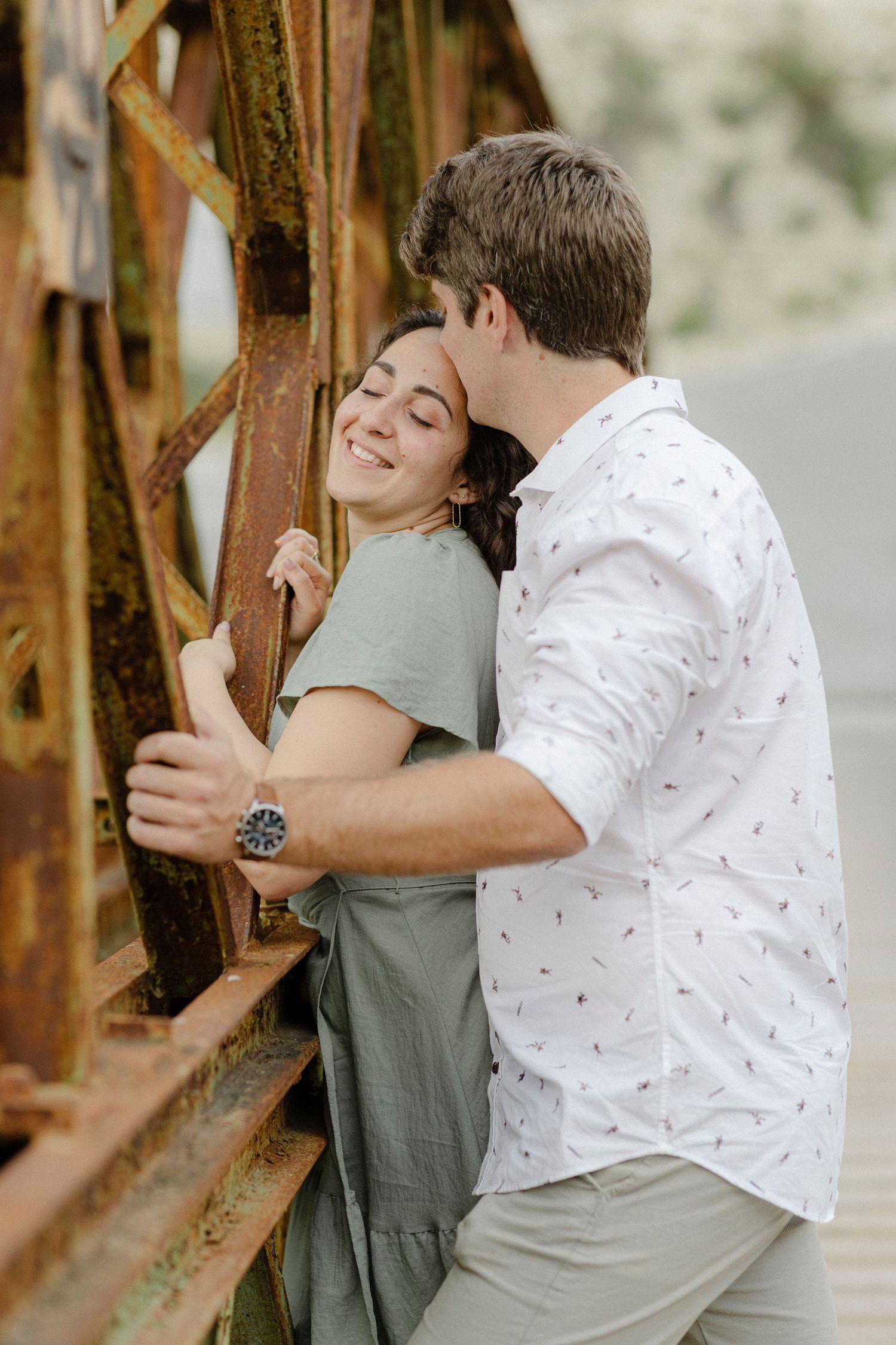Couple sharing a romantic moment against rusty metal structure, woman smiling with eyes closed as man kisses her forehead.