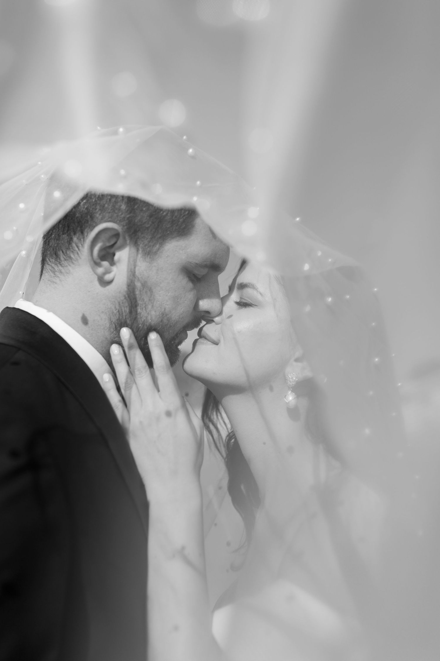 Romantic black and white photo of bride and groom kissing under a sheer veil with bokeh lights.
