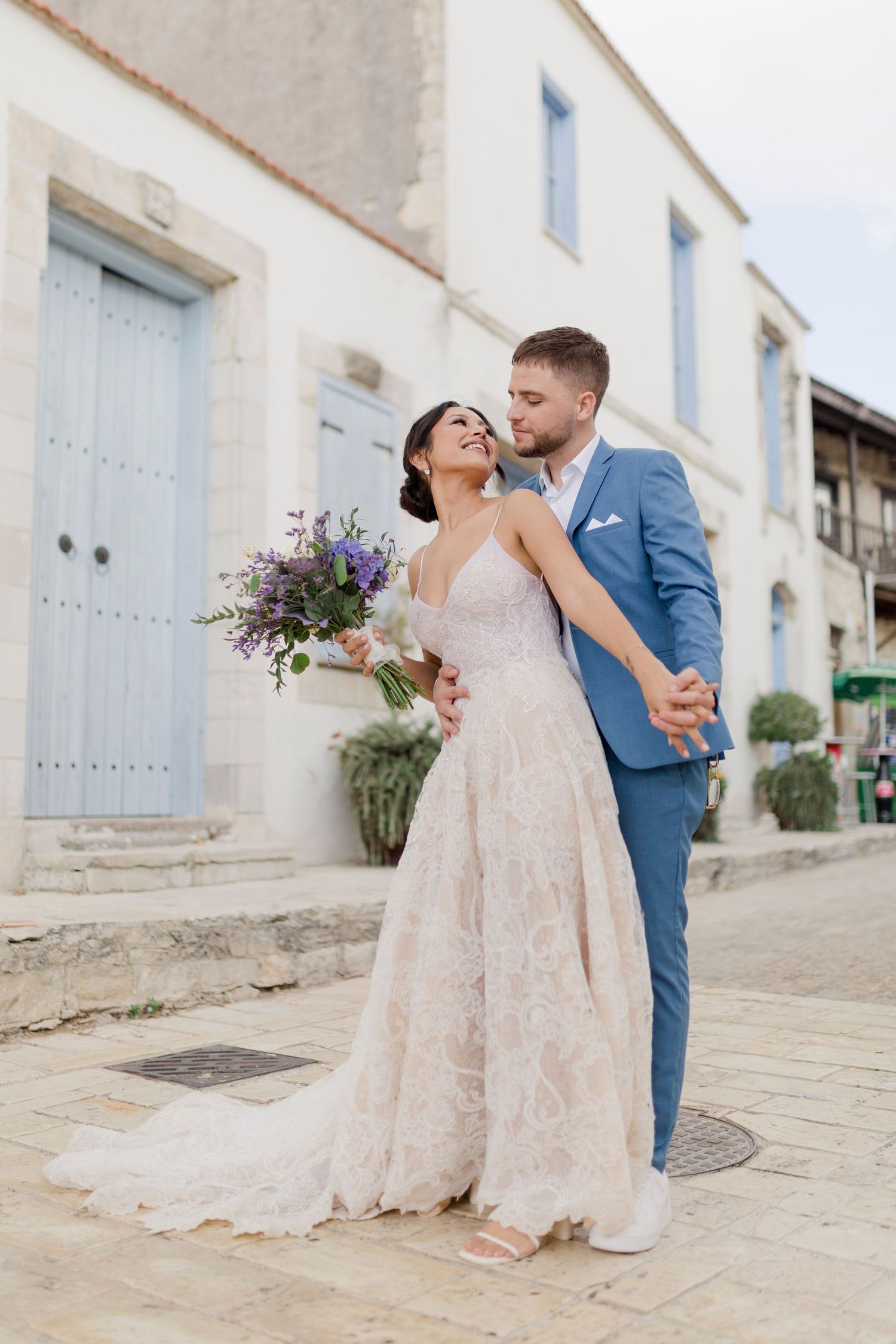 Bride in lace gown holds purple bouquet as groom in blue suit embraces her on cobblestone street.