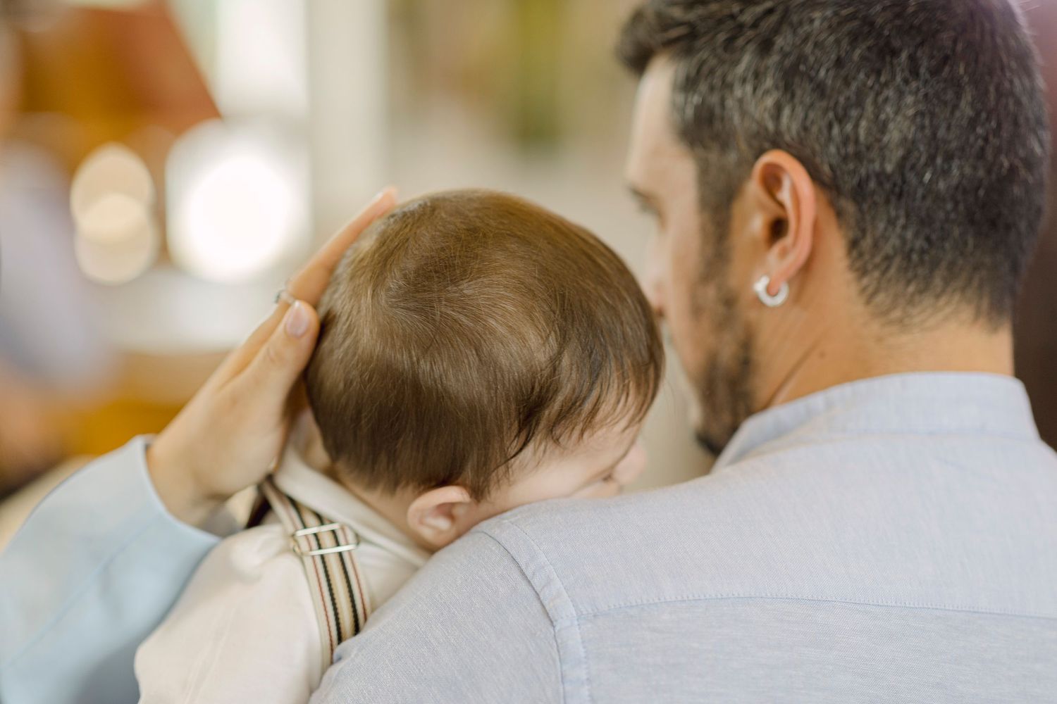 Father gently holding sleeping baby on his shoulder, viewed from behind in soft natural light.