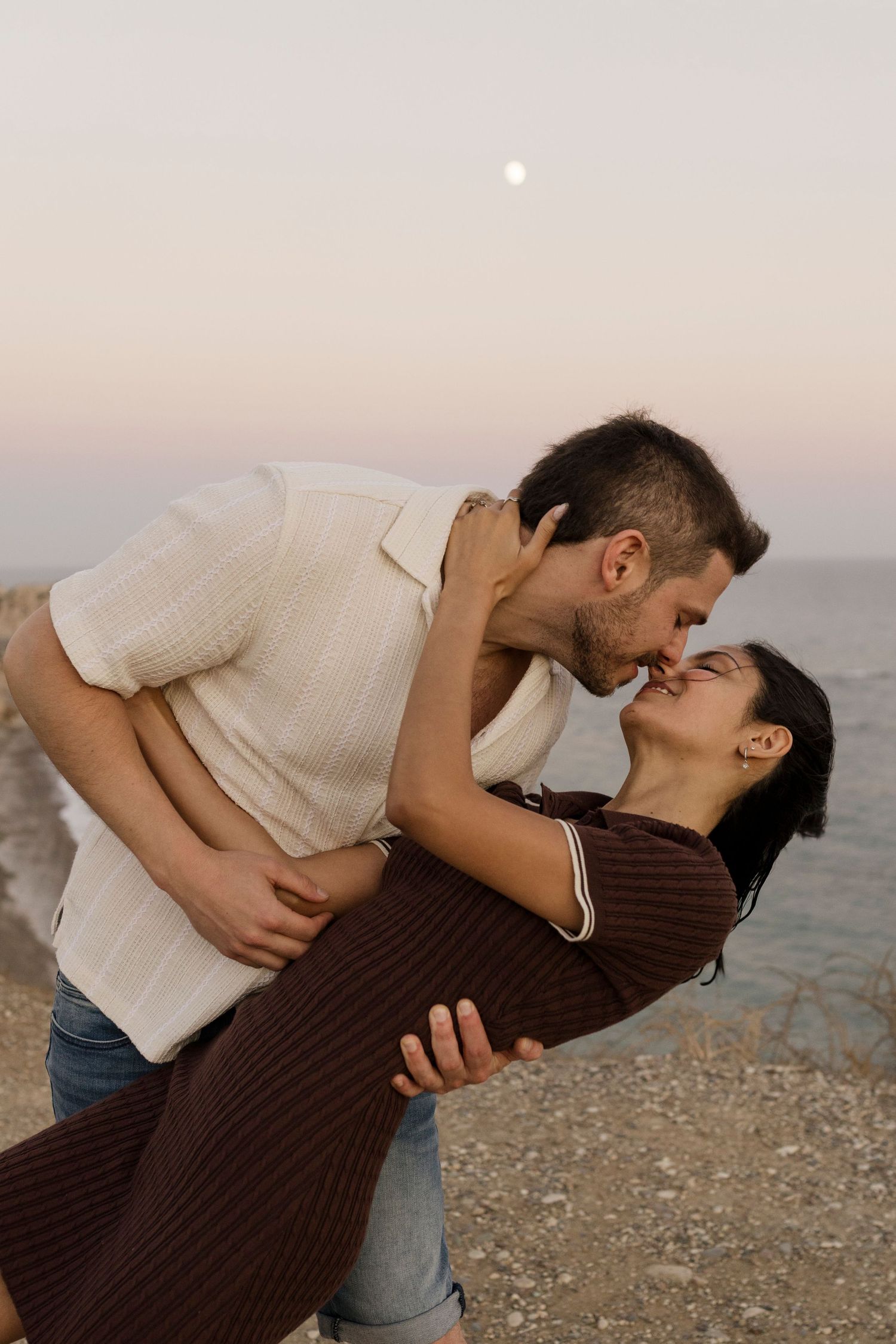 A couple shares a romantic kiss near the ocean at dusk with a moon visible in the pastel sky.