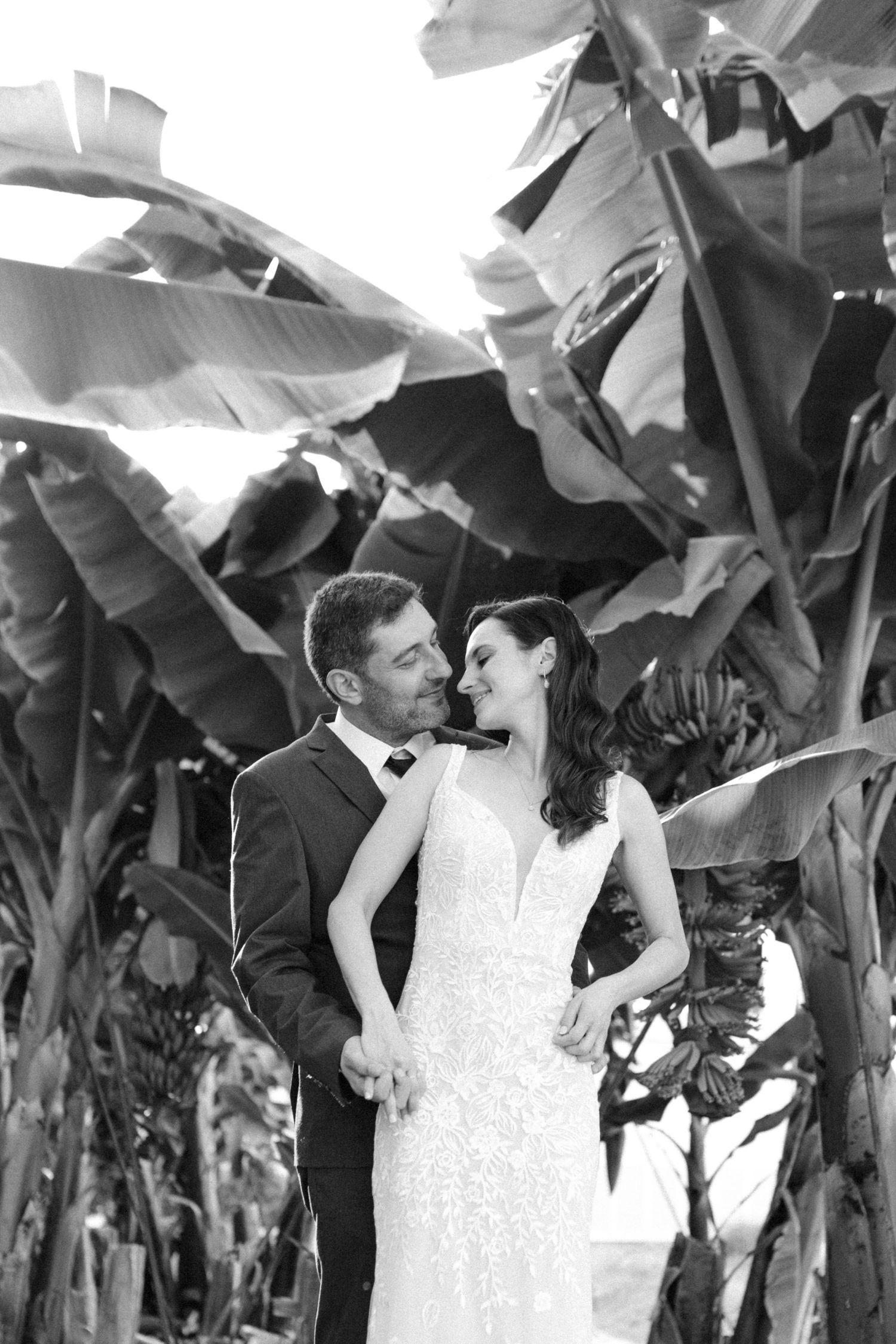 Black and white romantic portrait of bride and groom almost kissing among tropical banana tree leaves.
