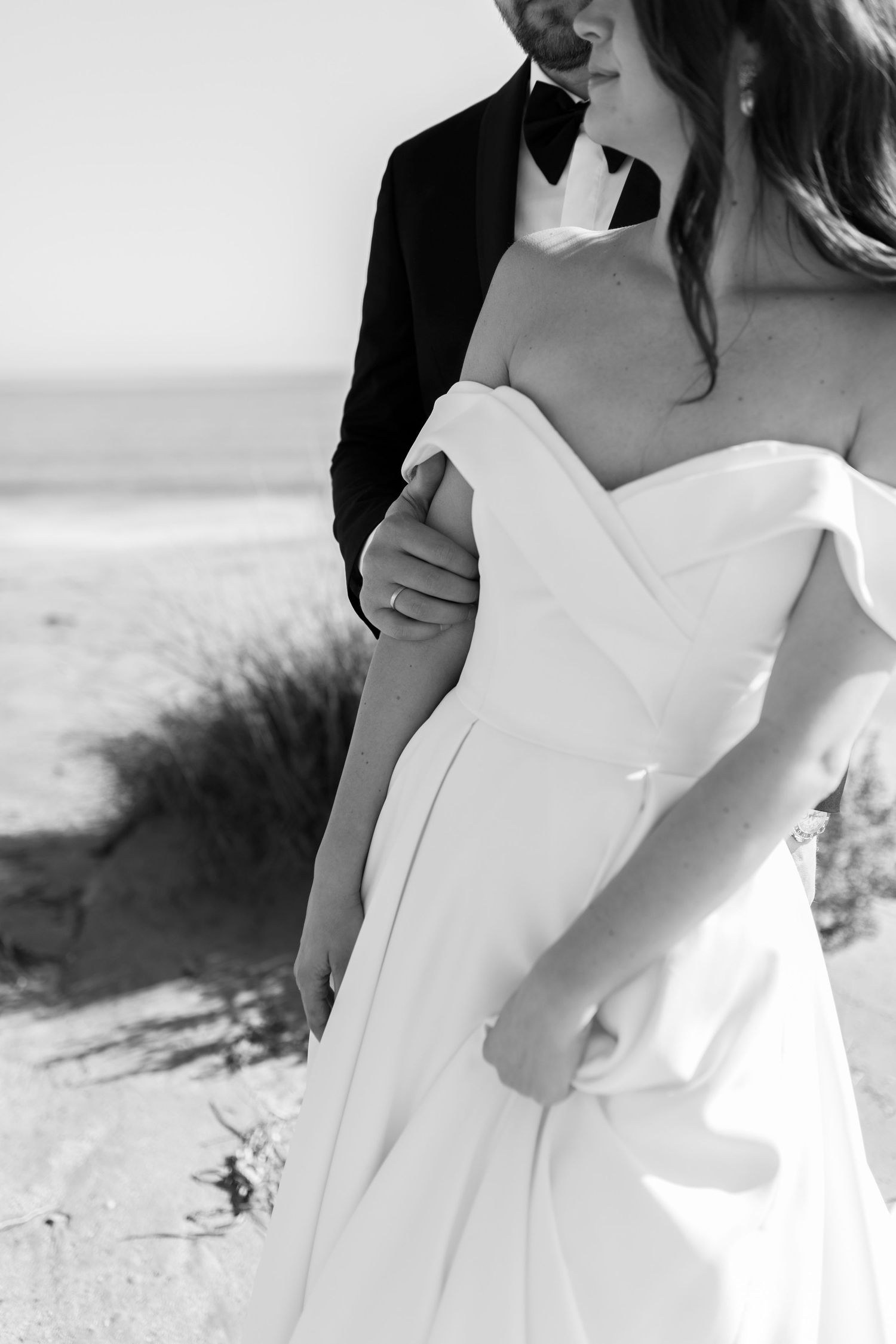 Black and white close-up of groom in tuxedo embracing bride in off-shoulder wedding gown on beach.