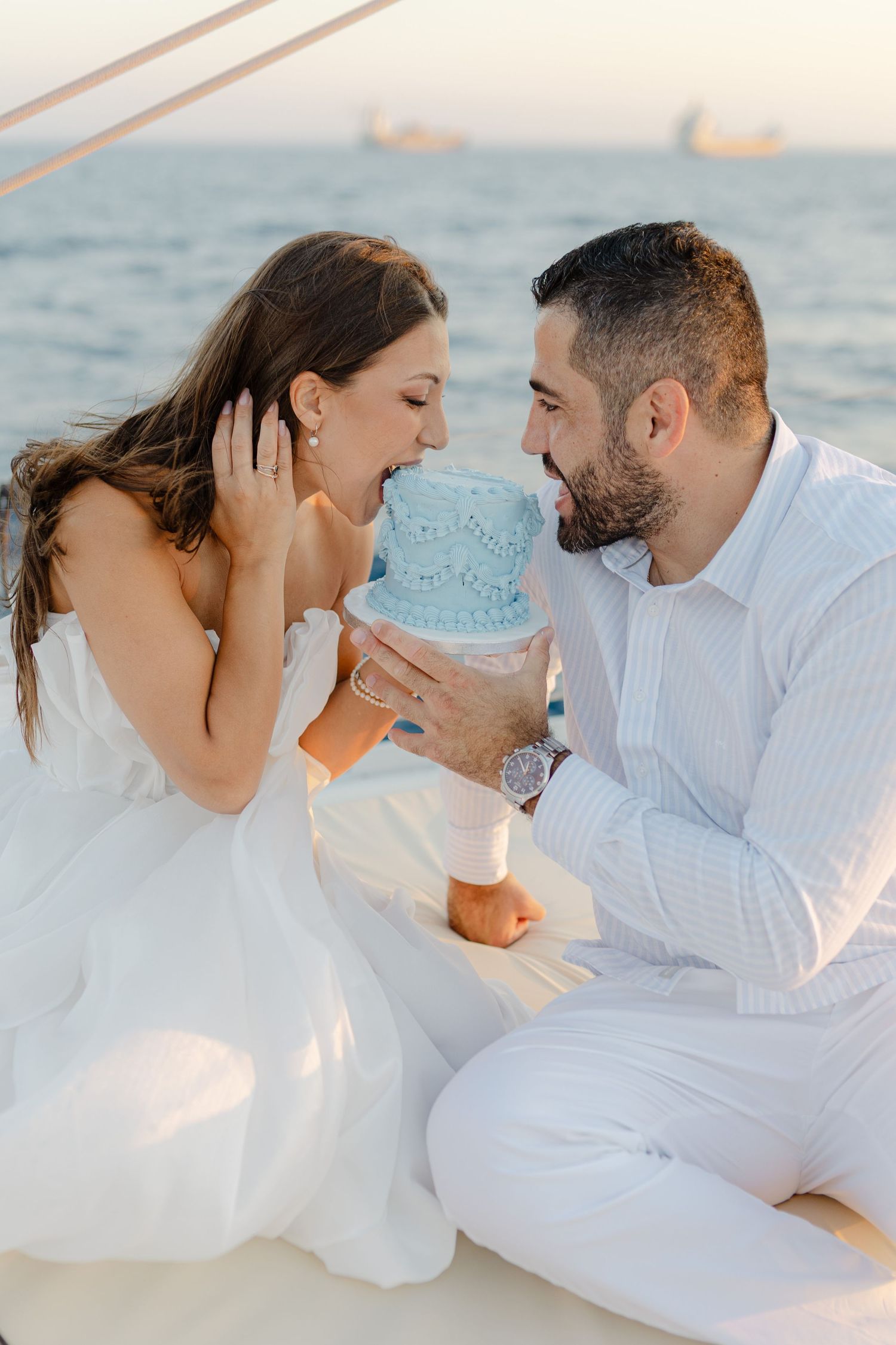Couple in white outfits sitting on a sailboat, sharing a joyful moment with a small blue cake by the sea.