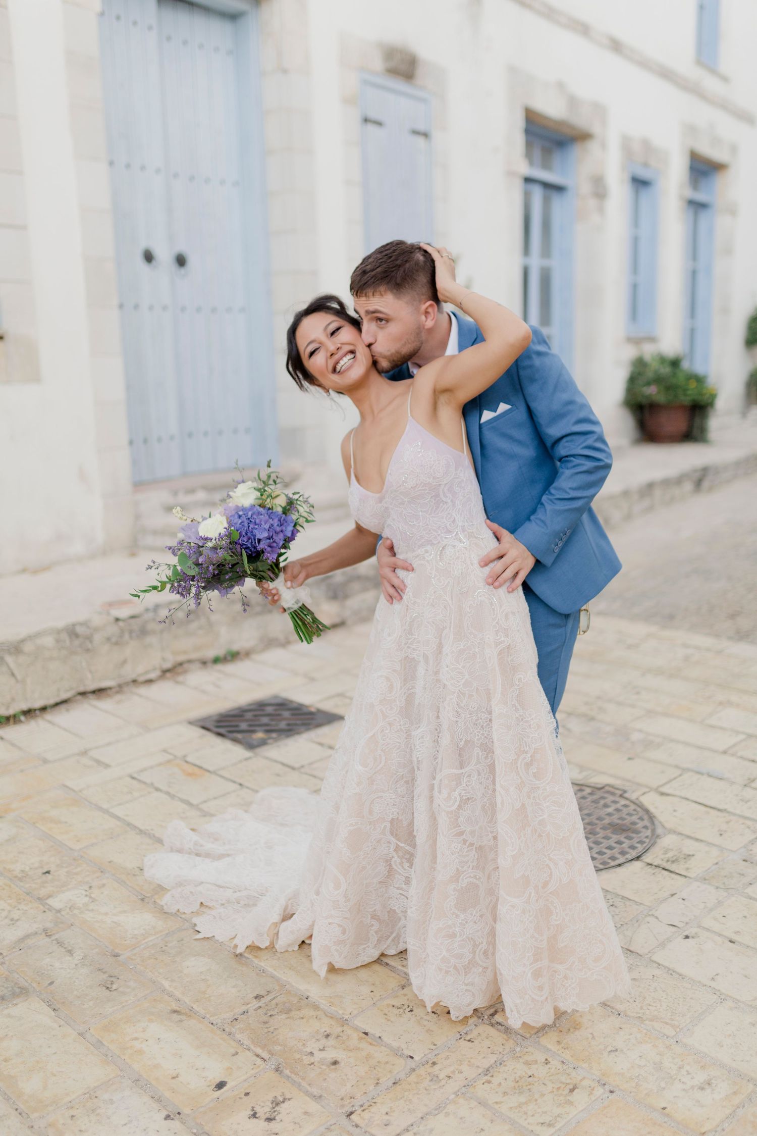 Bride in lace gown holds purple bouquet as groom in blue suit kisses her cheek on cobblestone street.