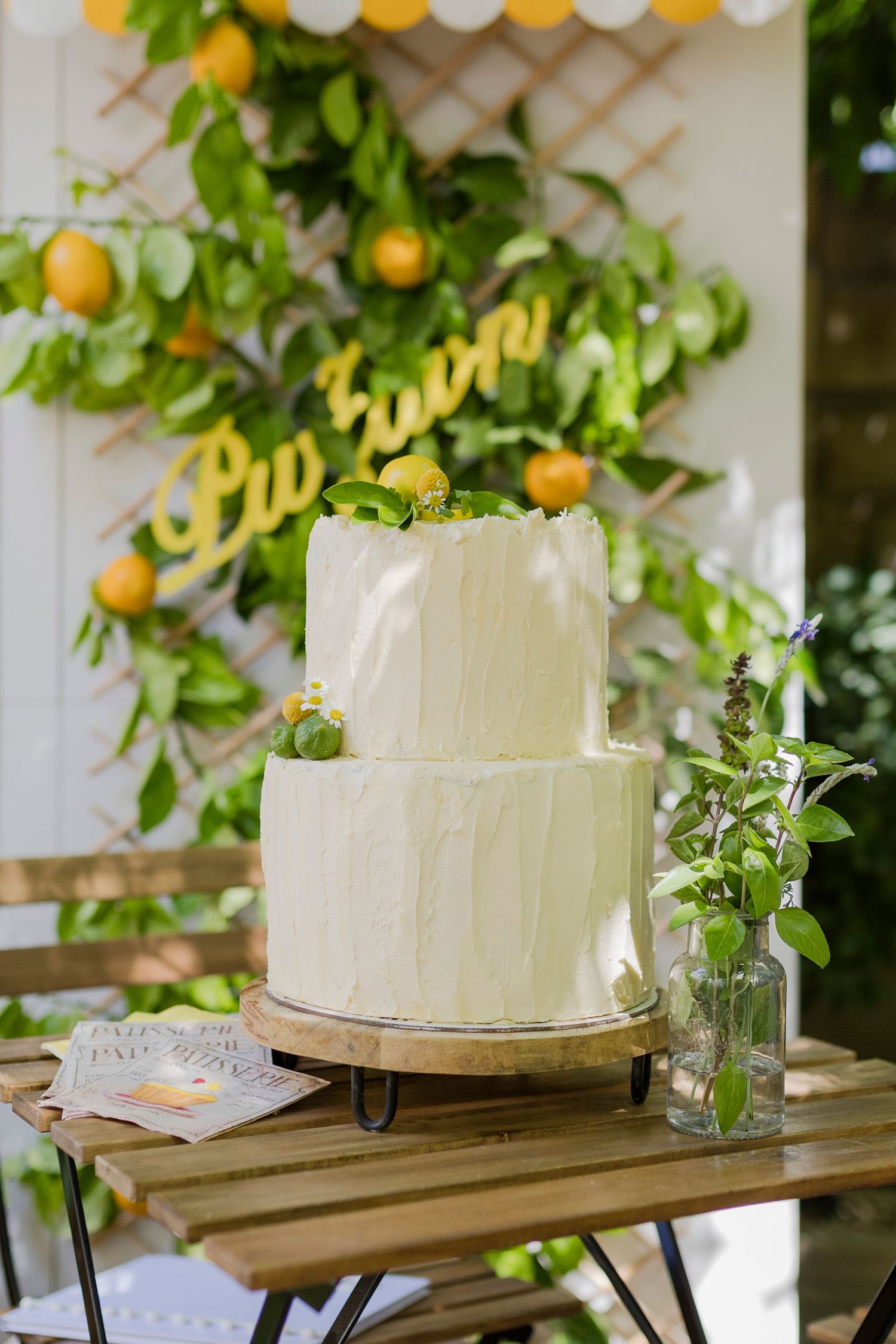 Two-tier white textured wedding cake with lemon decor on wooden table, green botanical backdrop with yellow citrus fruits.