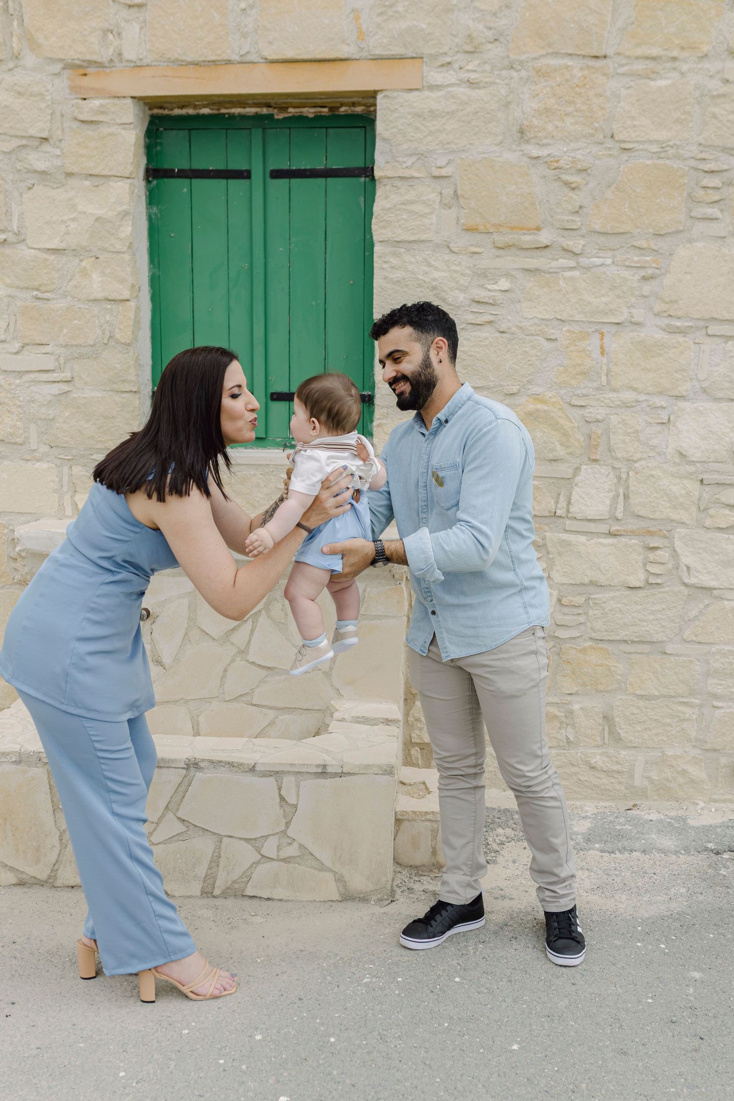 Happy couple in blue outfits playing with baby girl in front of stone wall with green shuttered window.