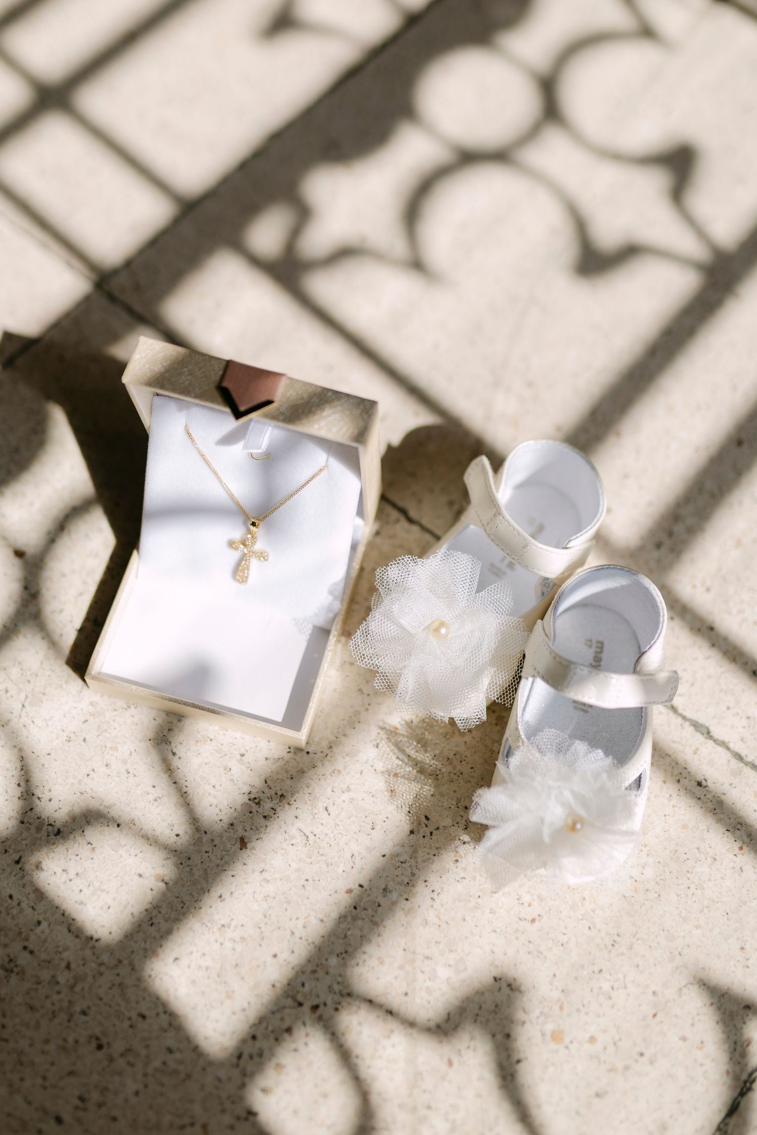 White baby shoes with tulle bows and gold cross necklace in box arranged on stone floor with decorative shadow pattern.
