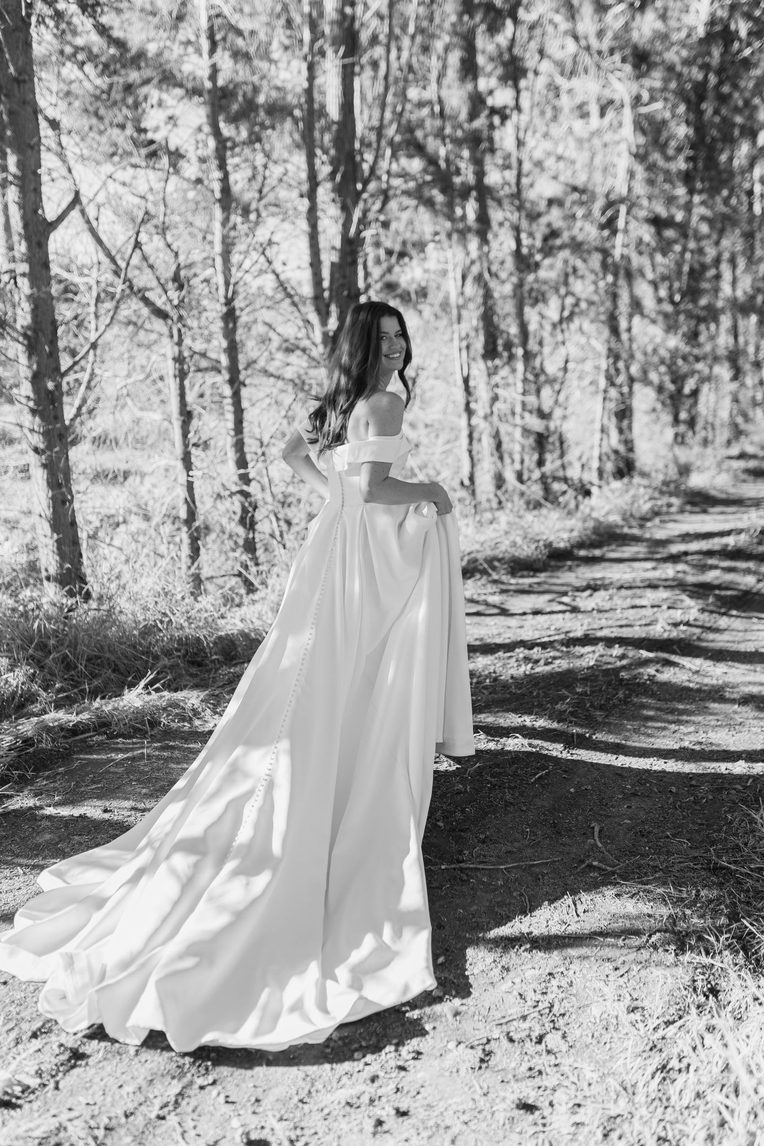 Black and white photo of bride in flowing off-shoulder wedding gown standing on sunlit forest path.