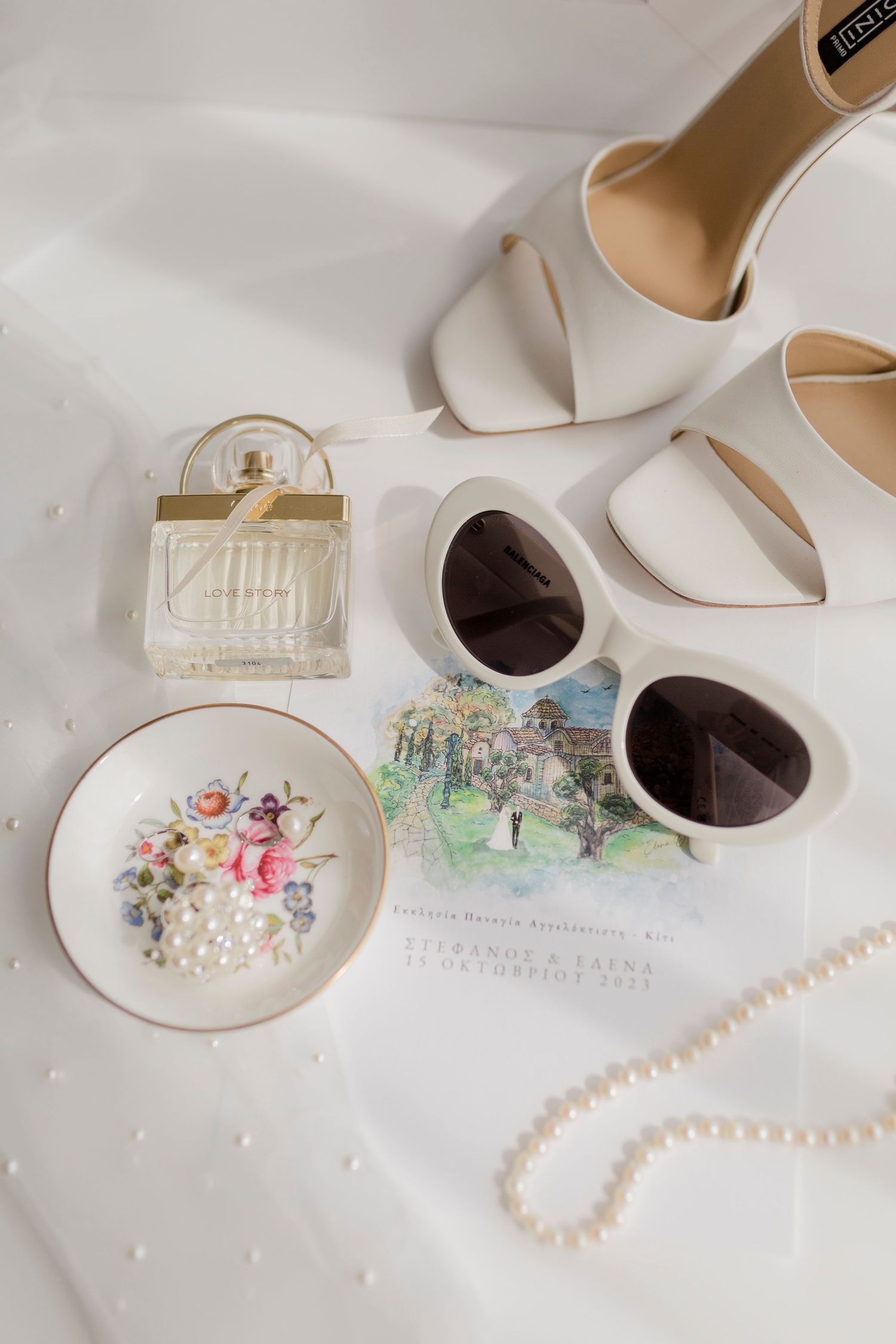 Elegant bridal flat lay with white heels, pearl necklace, sunglasses, floral dish, perfume, and wedding invitation on white fabric.