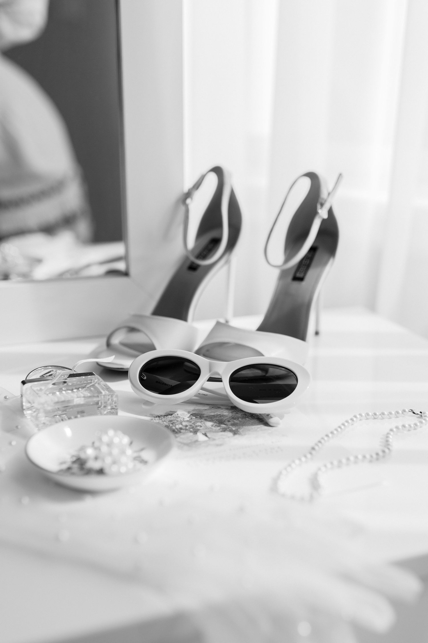 Black and white flat lay of bridal accessories including heels, sunglasses, pearl necklace, and perfume on white surface.