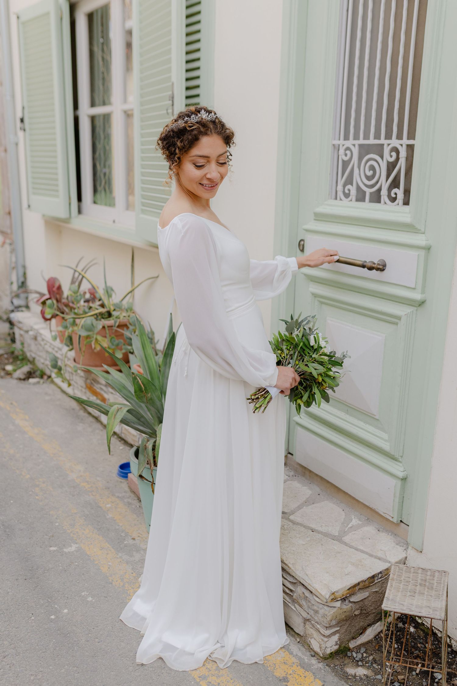 Bride in white long-sleeve gown holding green bouquet, knocking on a mint-colored door outdoors.