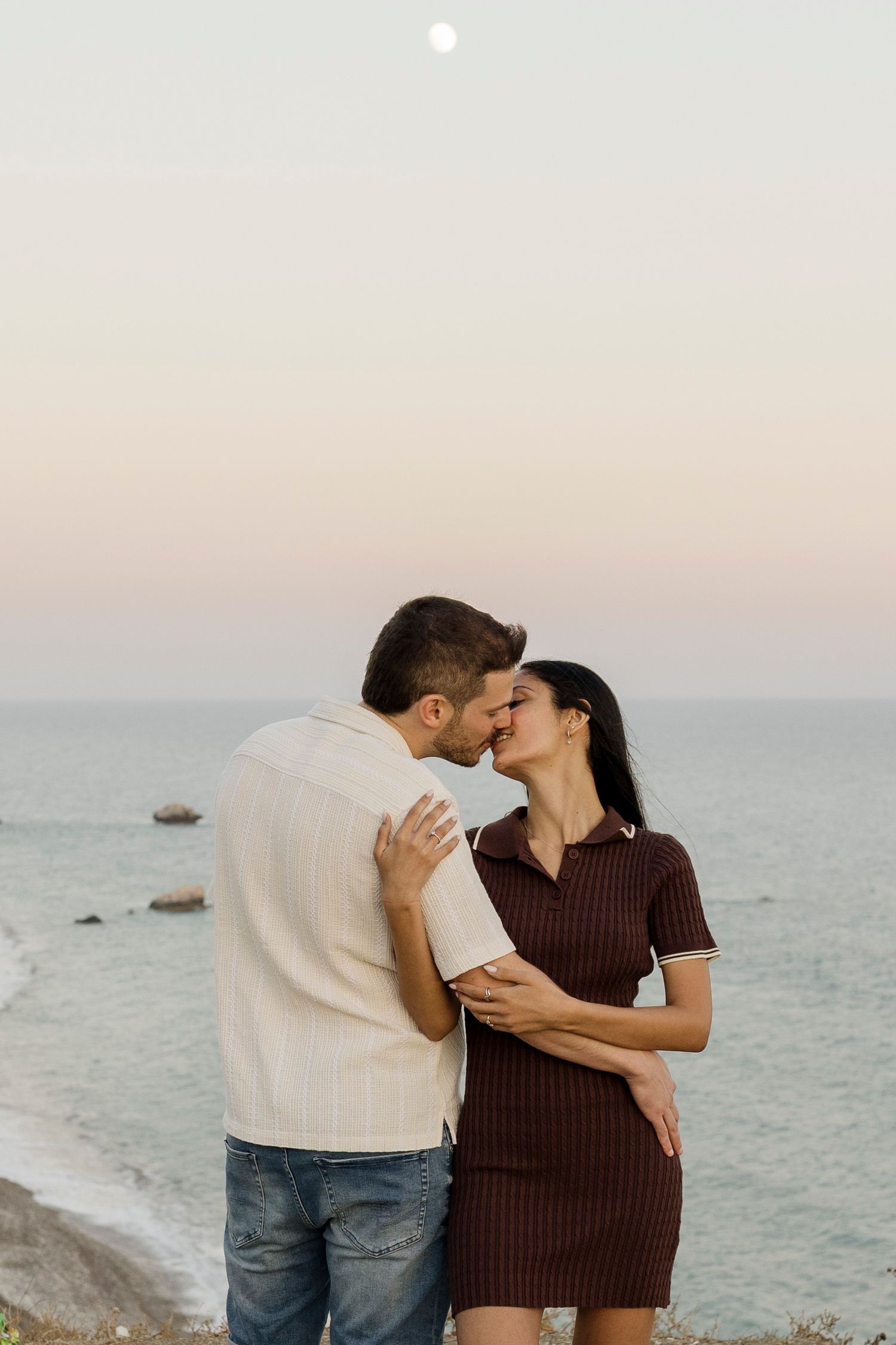 A couple sharing a romantic kiss by the ocean at dusk, with a full moon visible in the pastel sky.