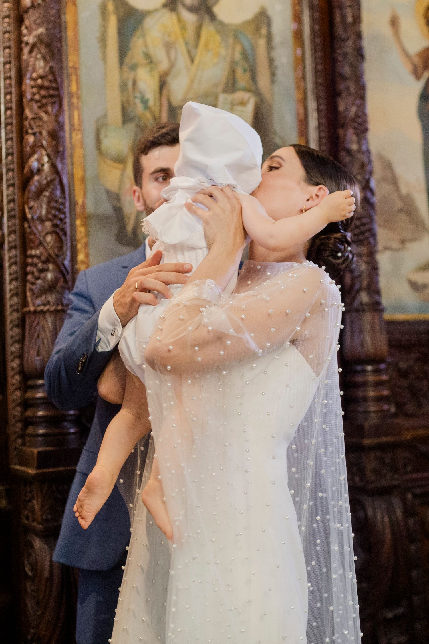 A mother in a white dress kisses her baby at a church baptism ceremony while the father stands behind them.